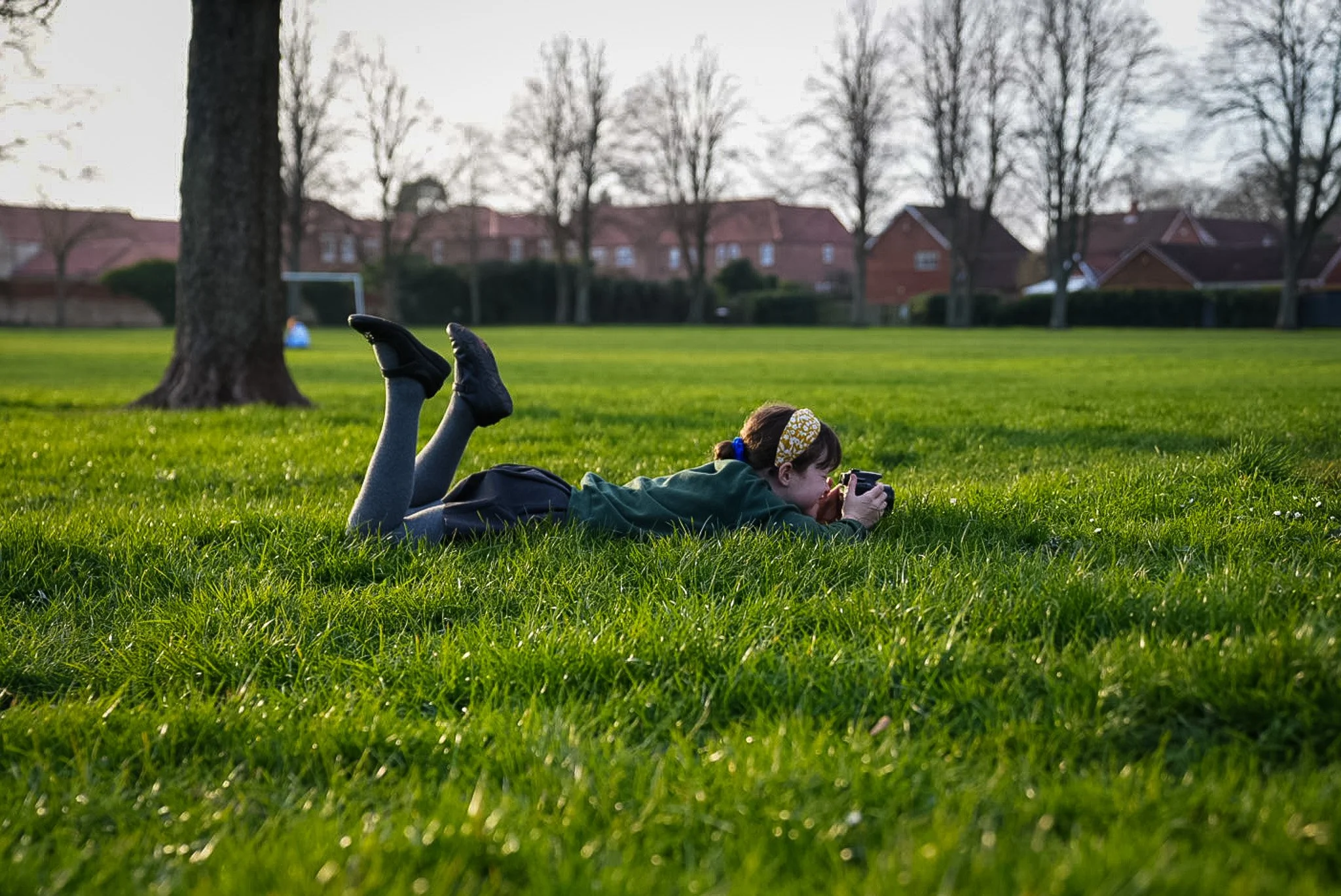 A girl lying on her stomach on green grass, taking a photograph with a camera. She is wearing a green jacket, gray tights, and black shoes, with a yellow headband. The background includes a tree, open field, houses, and a clear sky.