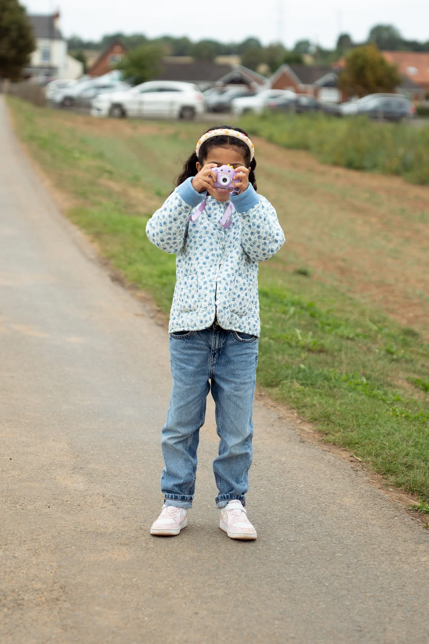 A young girl standing on a rural roadside, taking a picture with a purple instant camera. She is wearing a patterned jacket, jeans, and sneakers, with a headband, and there are cars and houses in the background.