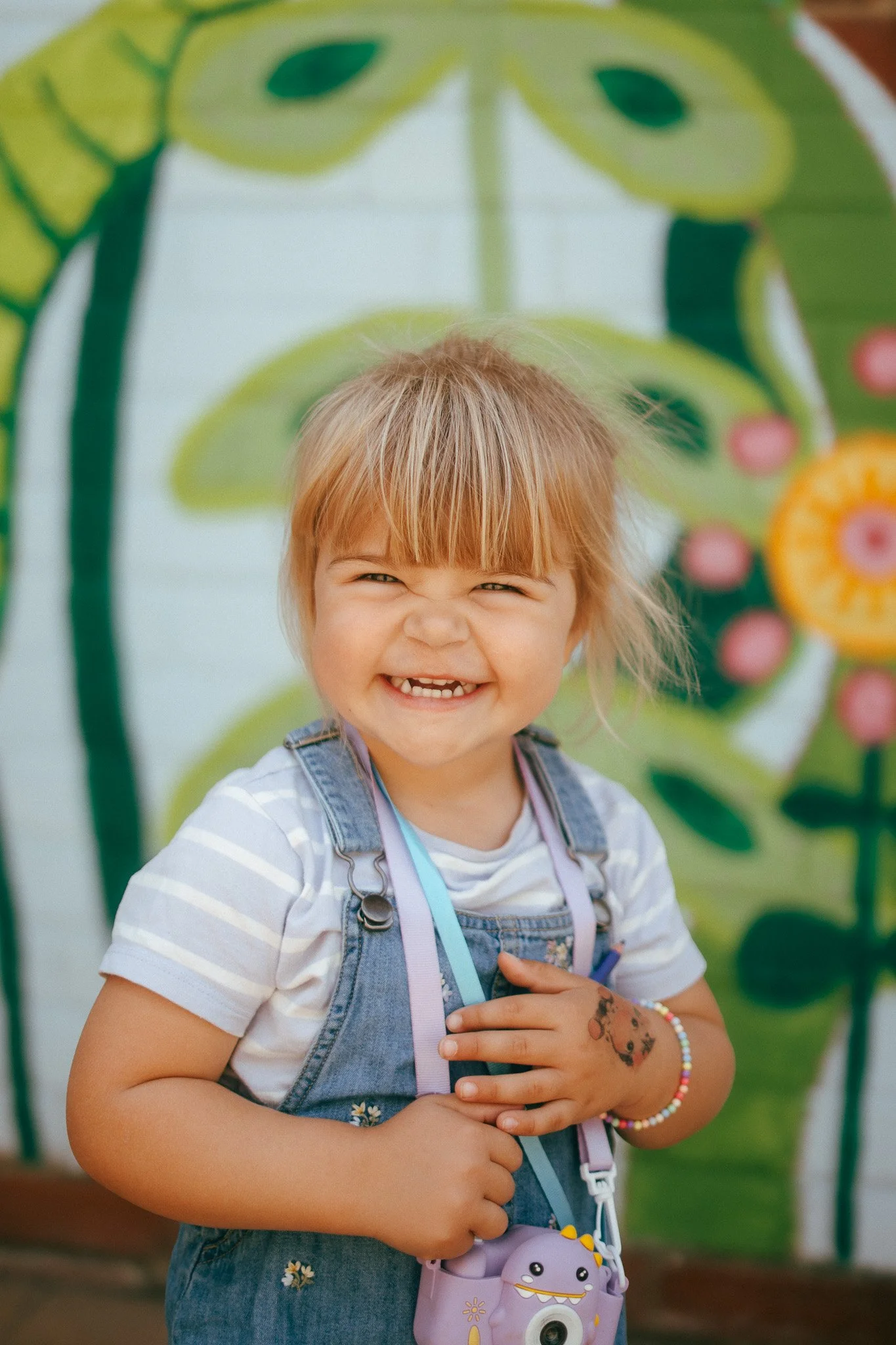 A young girl with red hair smiling and holding her hands on her chest, wearing a striped shirt, denim overalls, a colorful bracelet, and a camera around her neck, standing in front of a colorful painted background.
