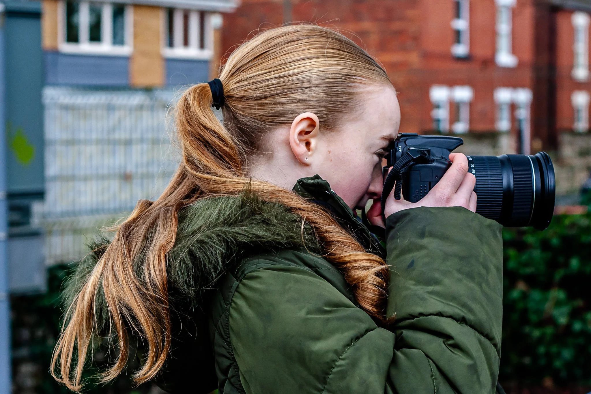 A young woman with red hair tied back in a ponytail, wearing a green jacket with a fur-lined hood, is looking through the viewfinder of a black digital camera outdoors in front of brick buildings and greenery.