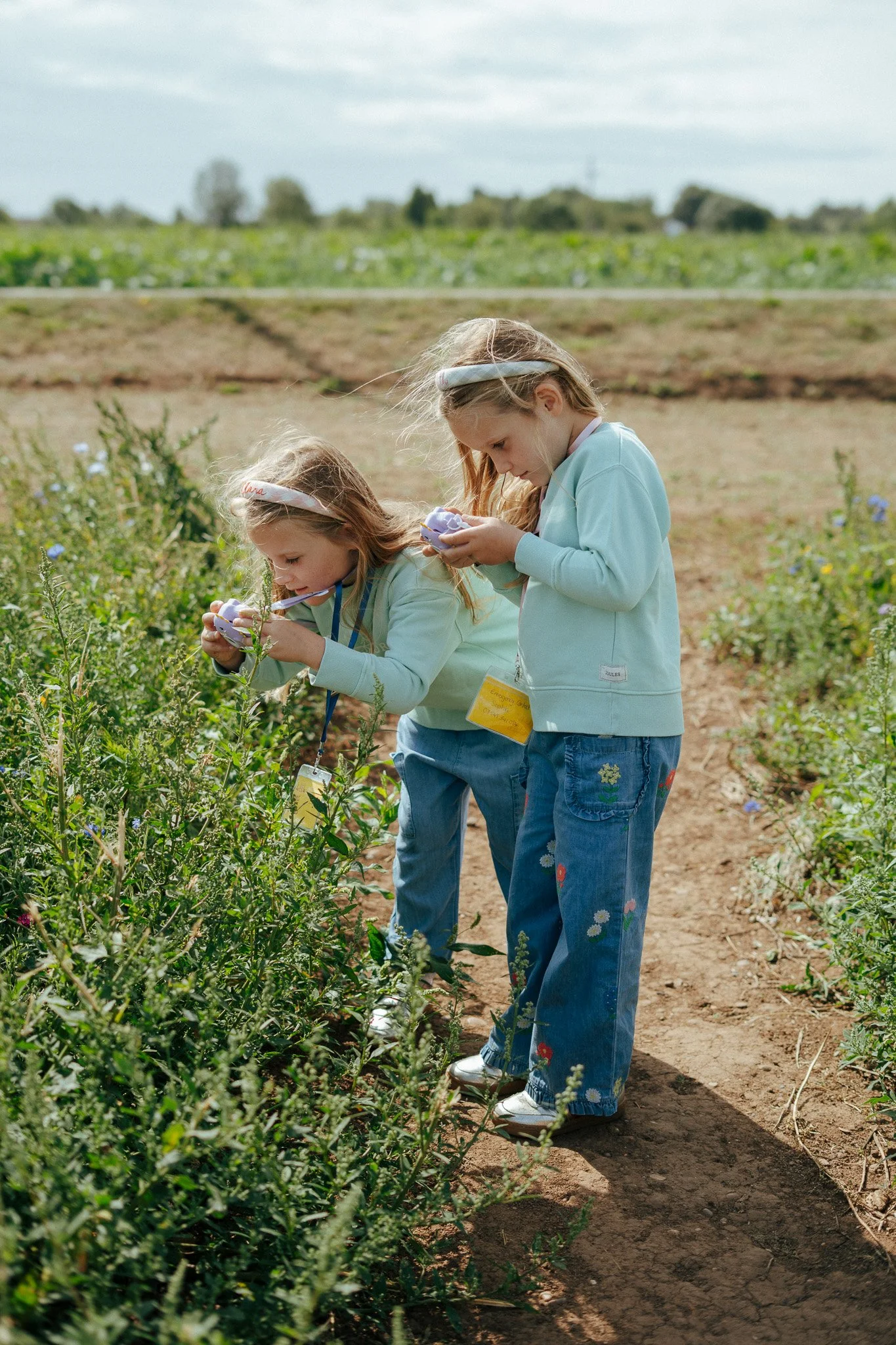 Two young girls with long hair, wearing light-colored sweatshirts and blue jeans with floral embroidery, are examining purple flowers in a field with green plants and a dirt path, under a cloudy sky.