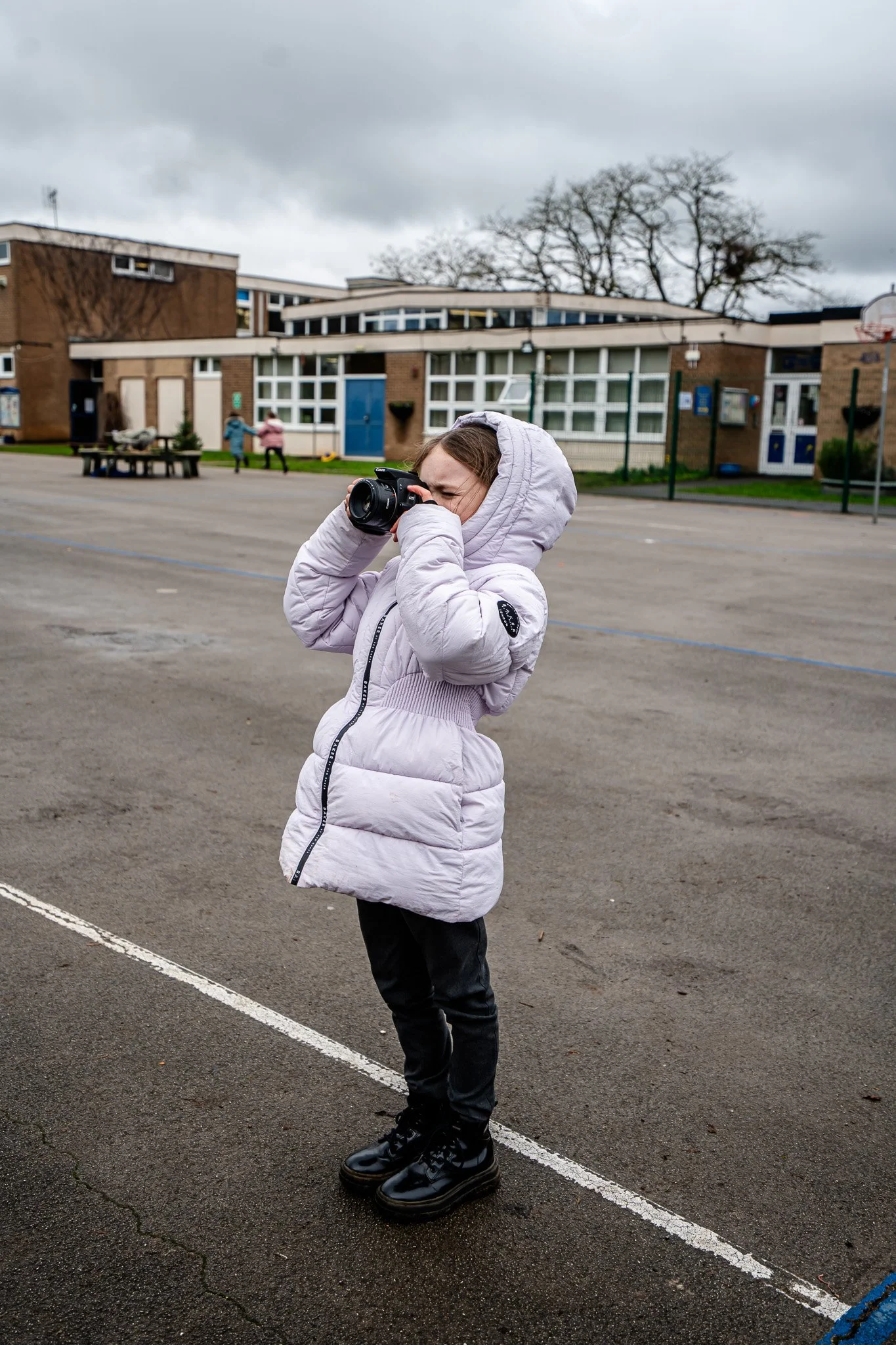 A young girl with a hooded pink jacket, black pants, and shiny black boots stands in an outdoor playground area, holding a camera up to her face and taking a photo. The background features a school building with large windows, a basketball hoop, and two children playing.