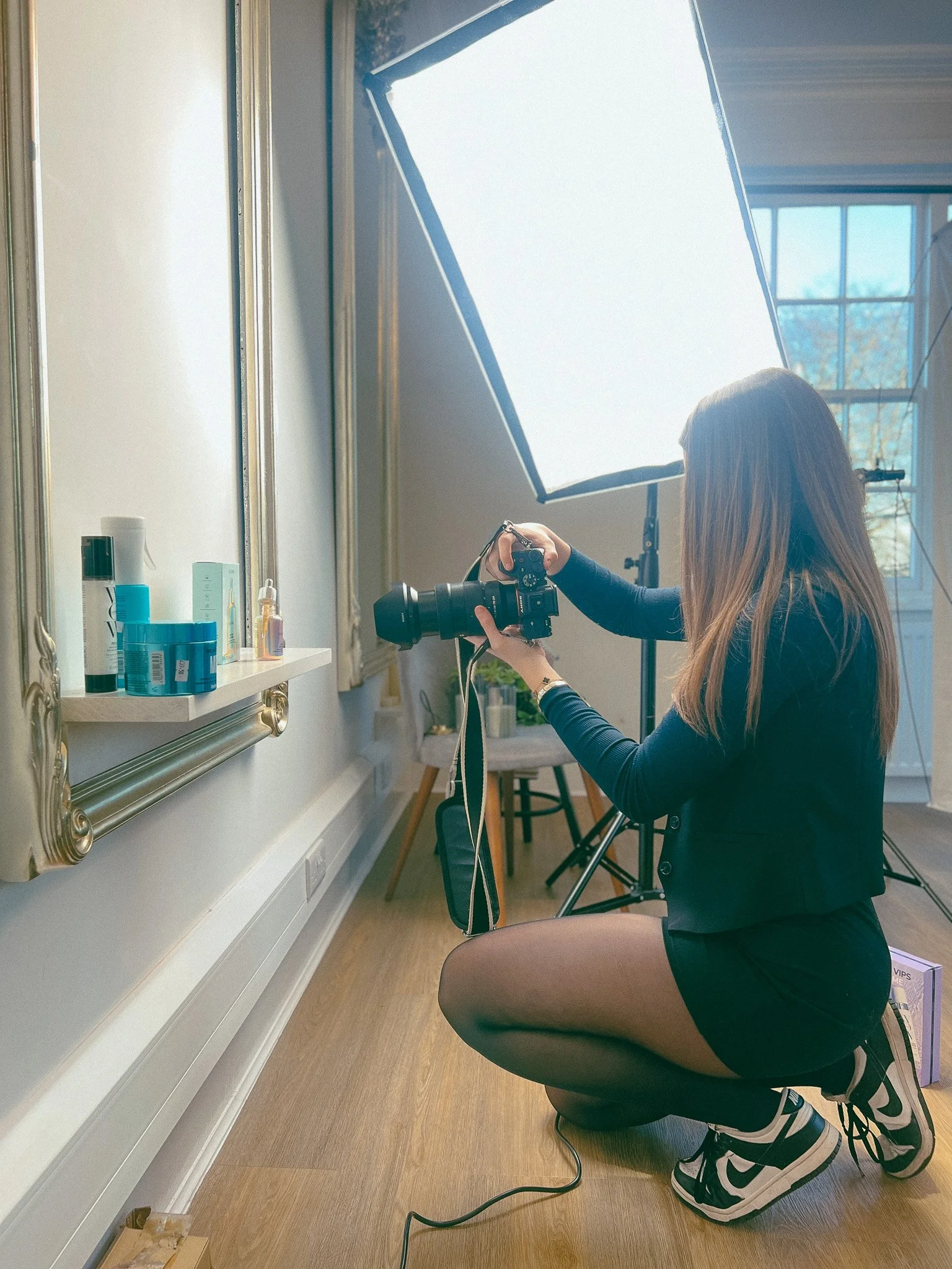 A woman kneeling on the floor in a photography studio, holding a camera, with a large softbox light behind her and a window in the background.