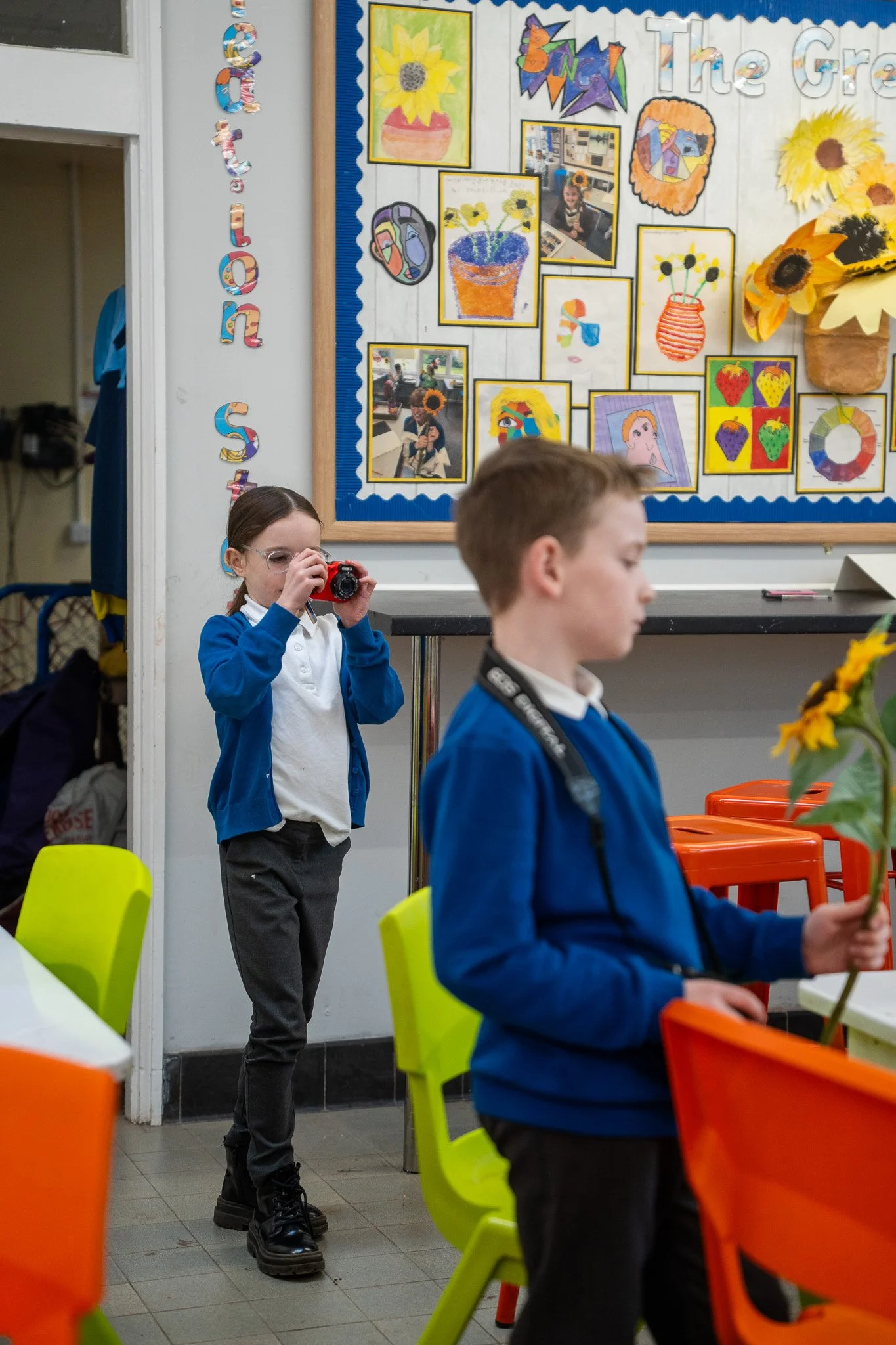 A girl in a blue school uniform taking a photograph with a red camera, standing behind a boy holding a sunflower, in a classroom decorated with colorful artwork depicting sunflowers and other bright colors.