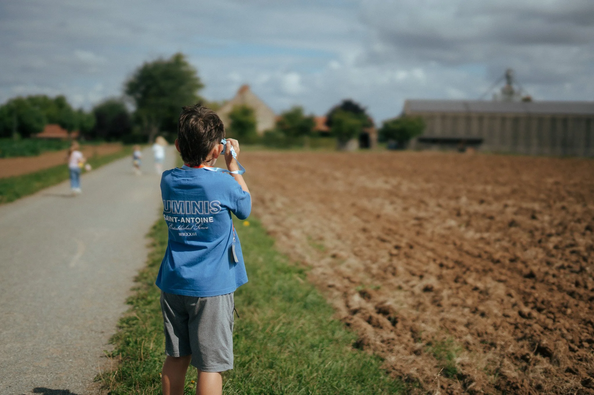 A young boy in a blue T-shirt and gray shorts taking a photo with a camera on a rural gravel path. Several other children are visible in the distance, walking or standing near a plowed field, with trees, farm buildings, and a cloudy sky in the background.