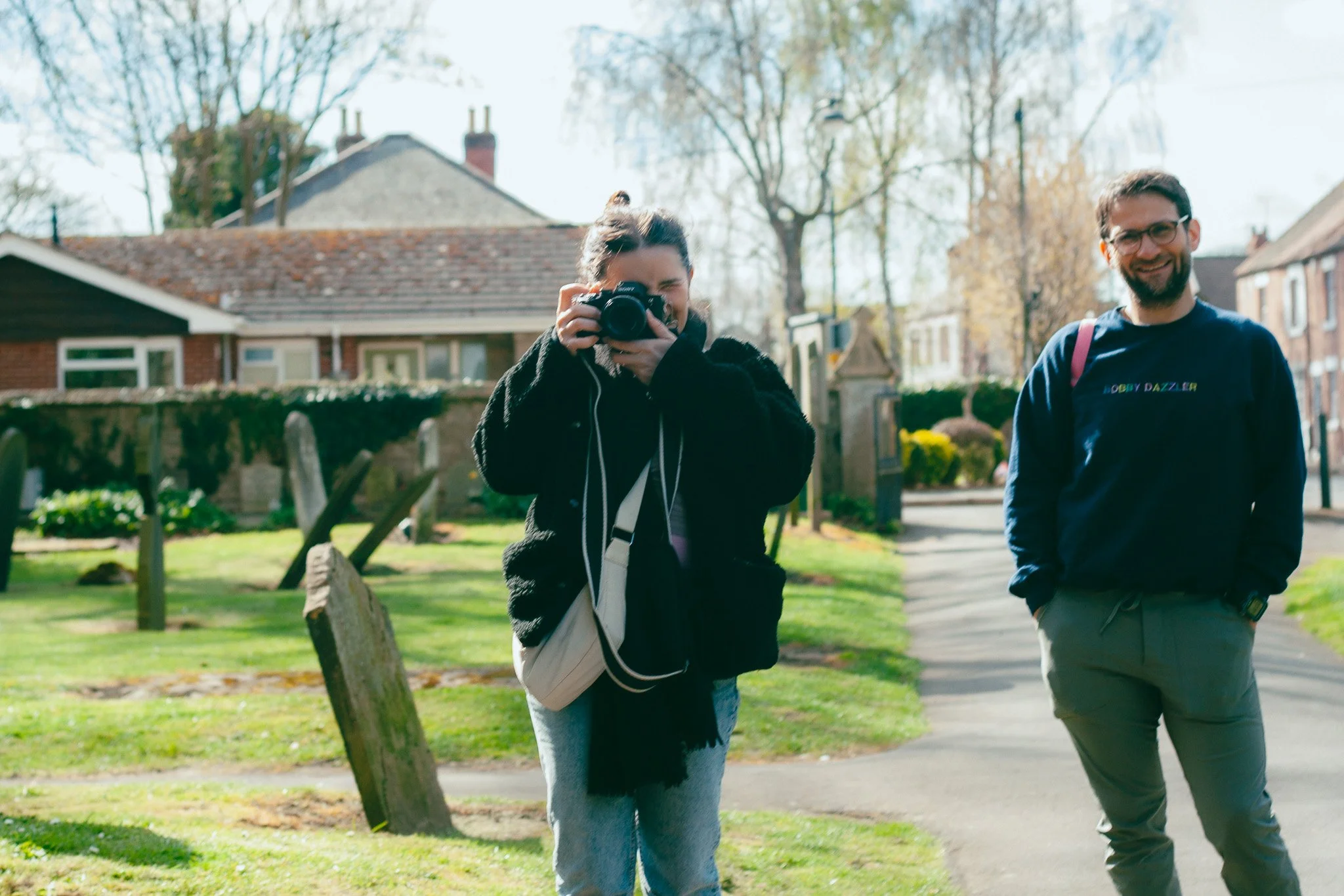 A woman taking a photo with a camera and a man smiling nearby in a park on a sunny day.
