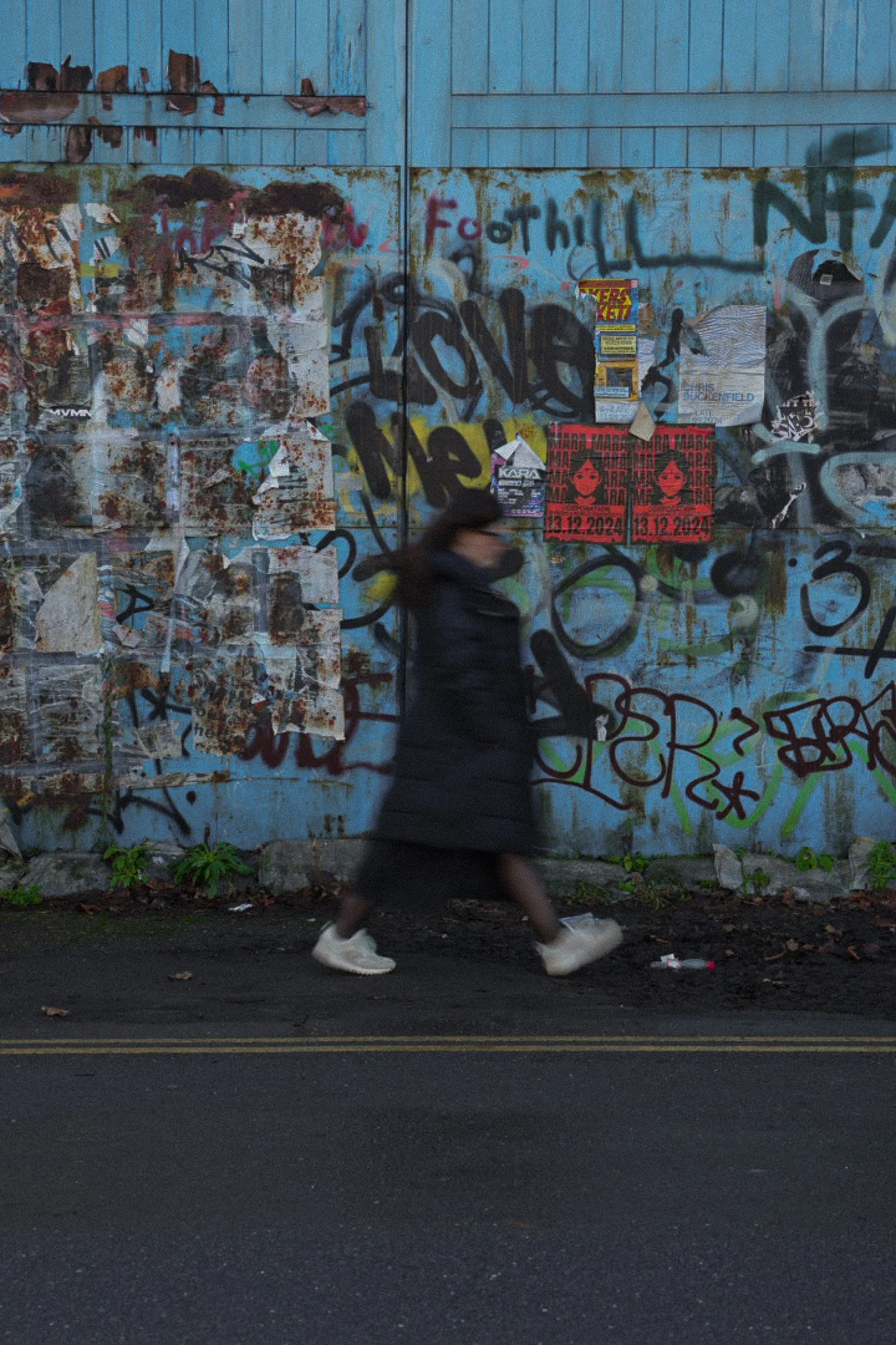 A woman walking briskly past a graffiti-covered blue wall with posters, on a street with dark pavement and a double yellow line.
