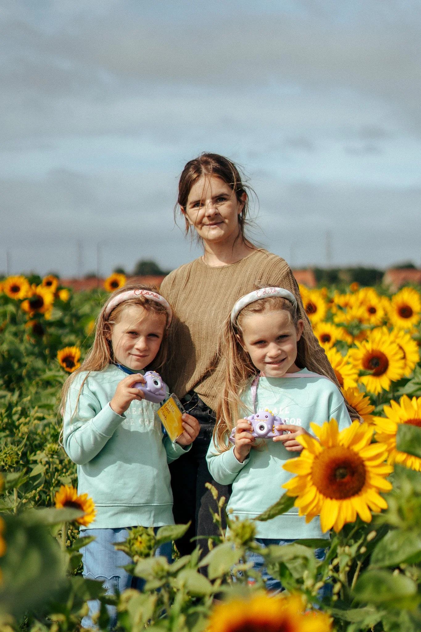A woman and two young girls standing in a sunflower field, holding cameras and smiling at the camera.