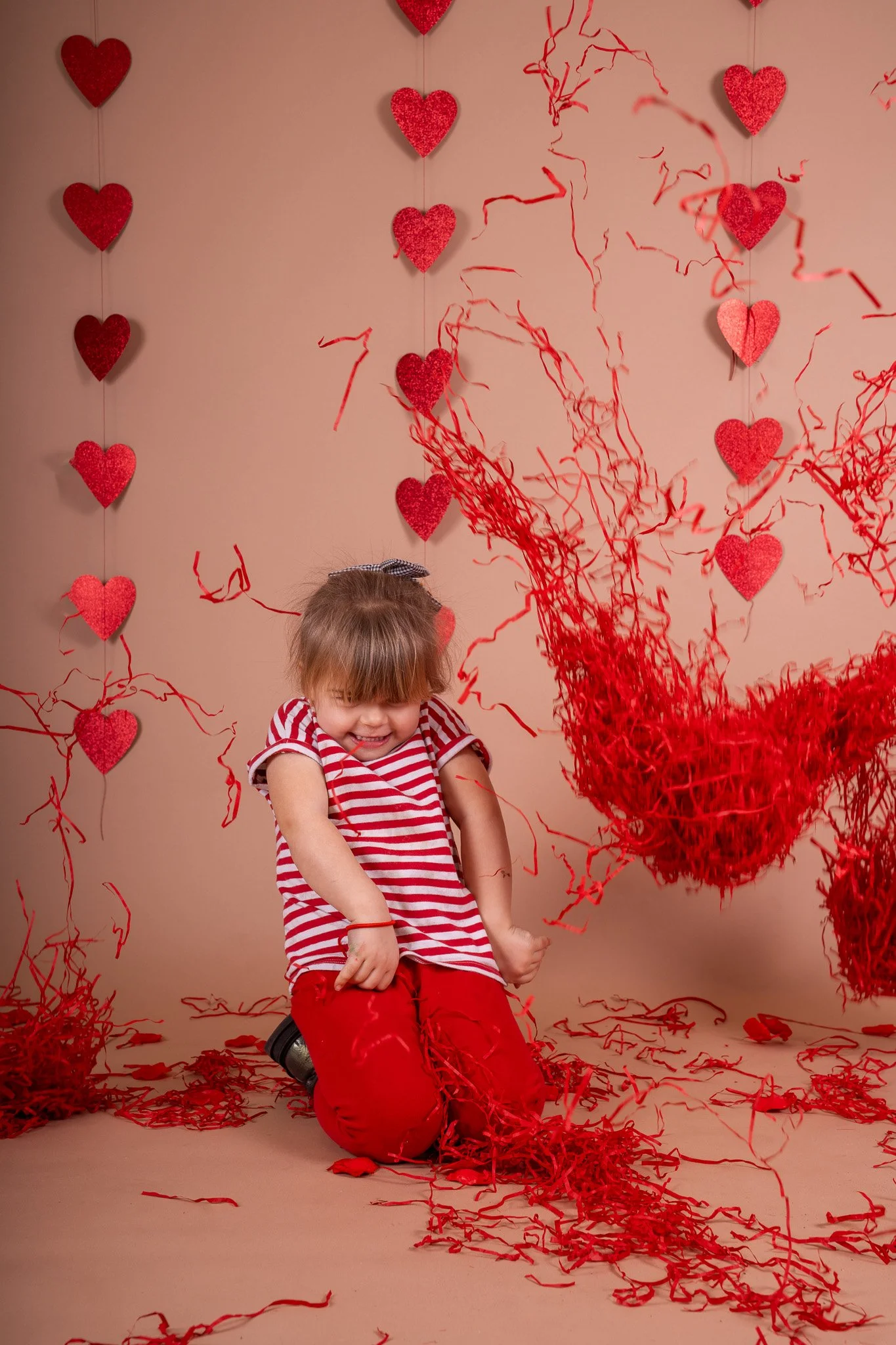 A young girl in a red and white striped shirt and red pants kneeling on the floor with red paper streamers around her, surrounded by Love-themed decorations including red hearts hanging on the wall and red streamers, celebrating Valentine's Day.
