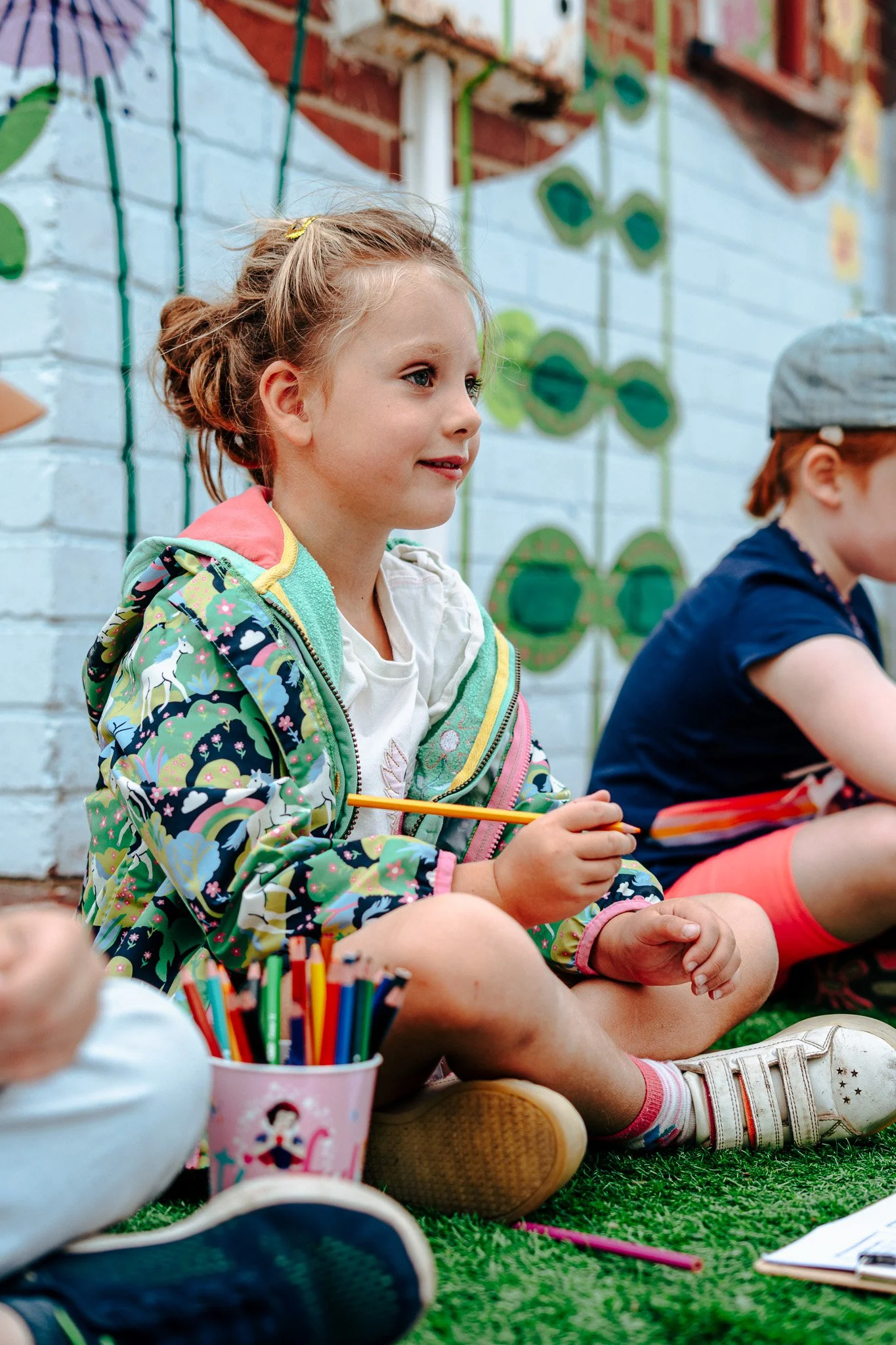 Young girl sitting on grass with a colorful jacket, holding a yellow pencil, and surrounded by colored markers and paper, engaging in an outdoor activity with other children nearby.