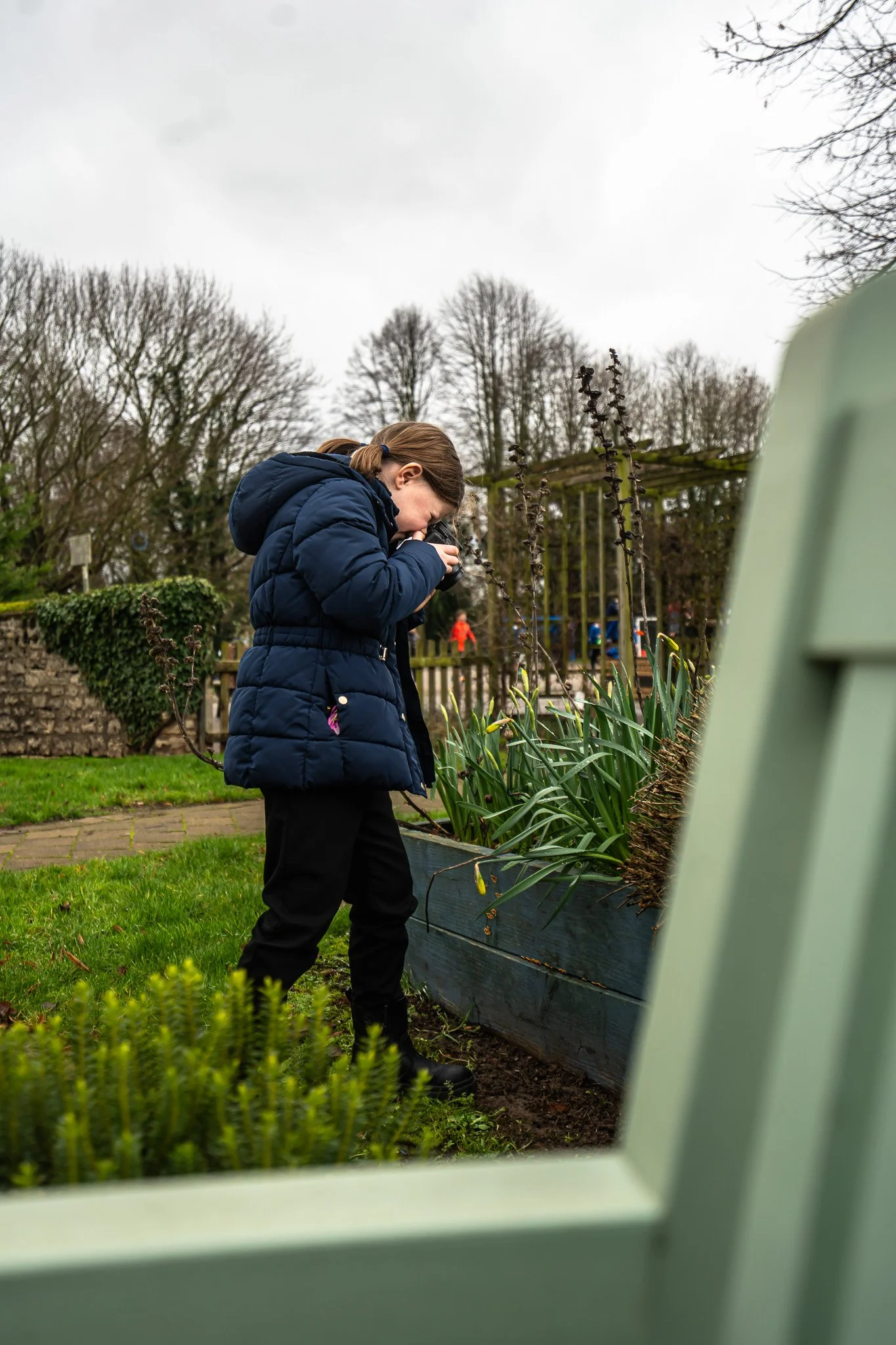 A girl in a blue puffer jacket and black pants looking closely at plants in a garden bed through a magnifying glass, with a green chair partially visible in the foreground.