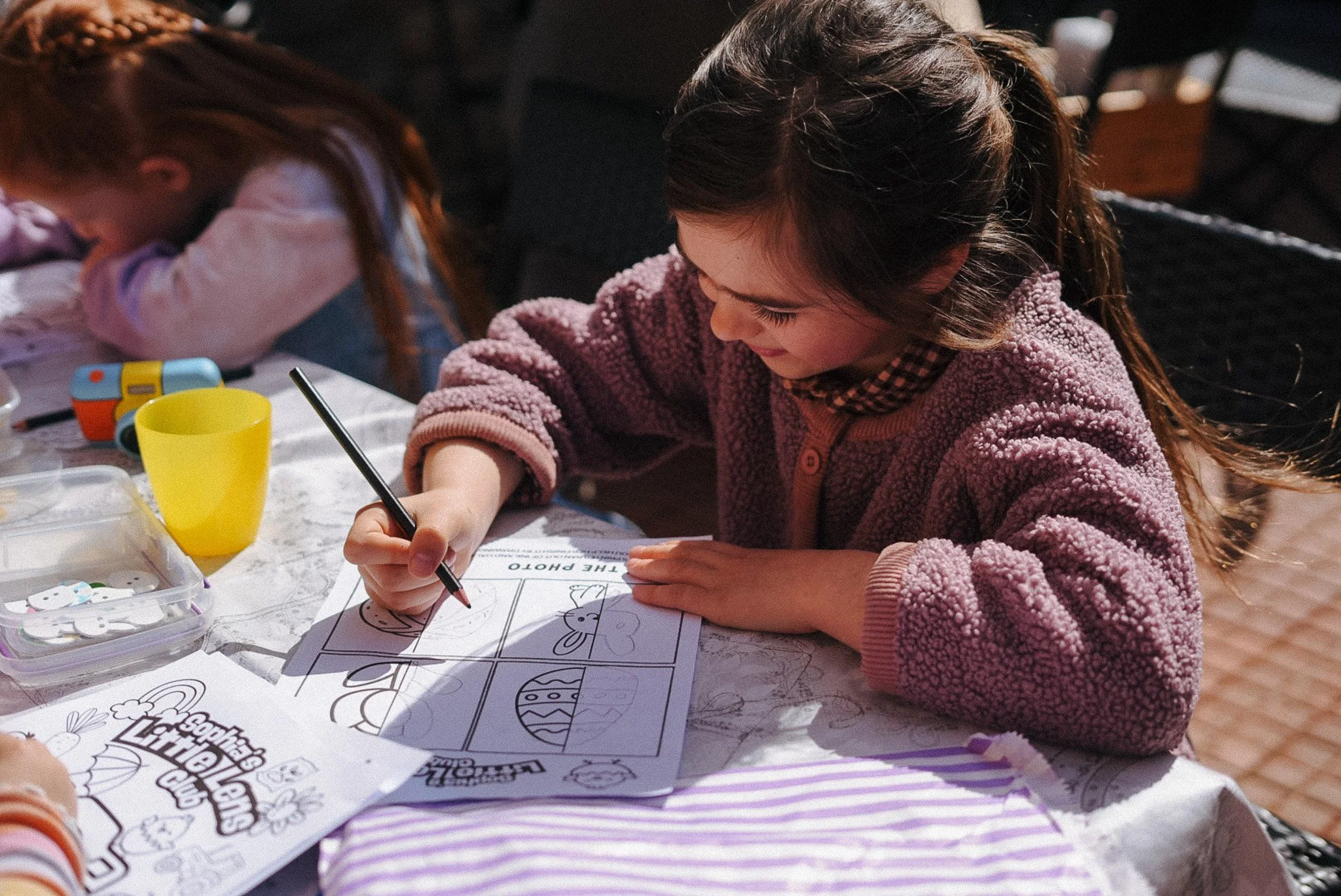 A young girl is coloring in a Easter-themed activity book at a table, with another child partially visible beside her. The table has coloring supplies, including markers, a yellow cup, and a box of stickers. The girl is focused on her coloring, wearing a pink fuzzy jacket.