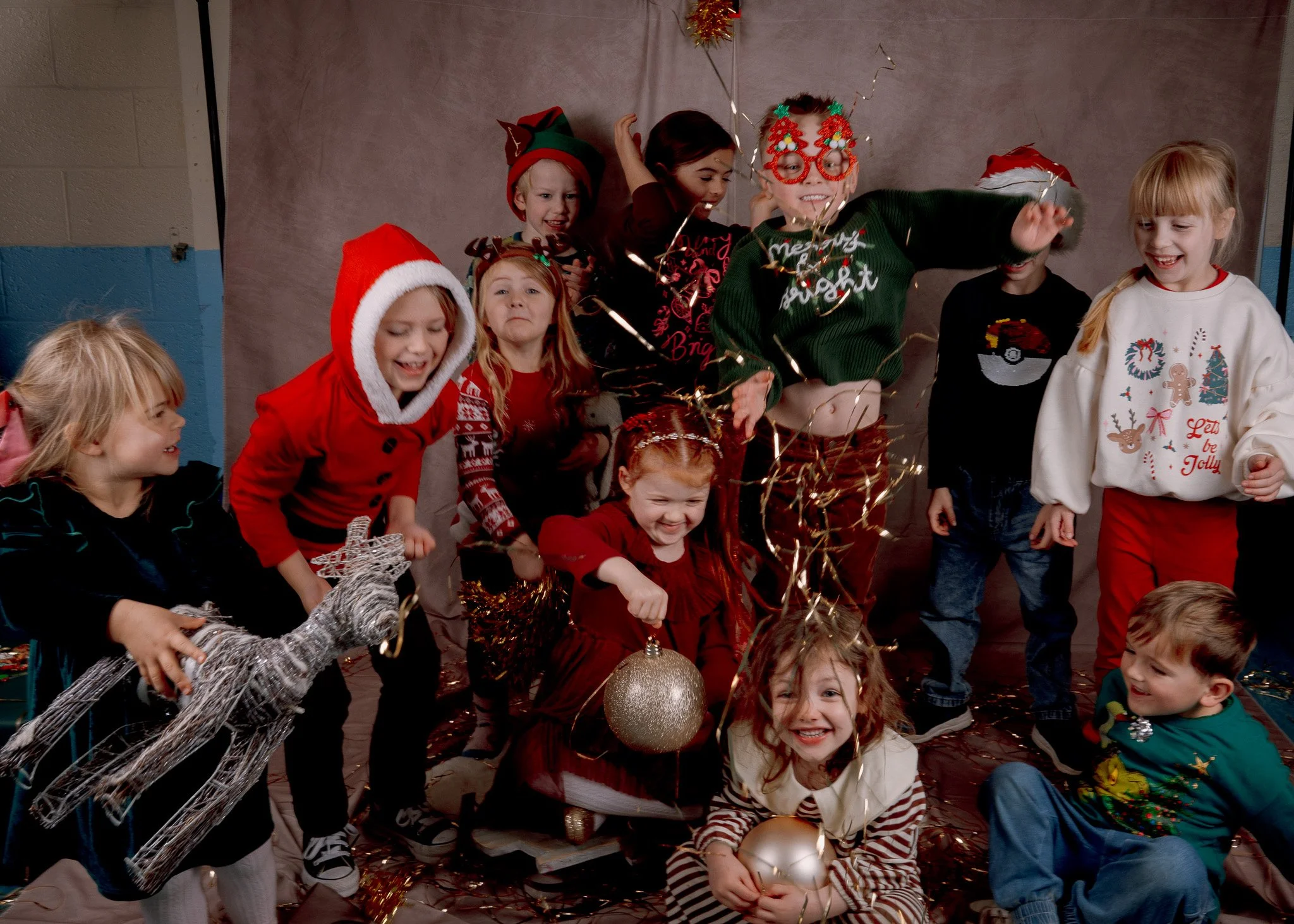 Children in festive Christmas sweaters and costumes celebrating together, holding ornaments, surrounded by tinsel and sparklers.
