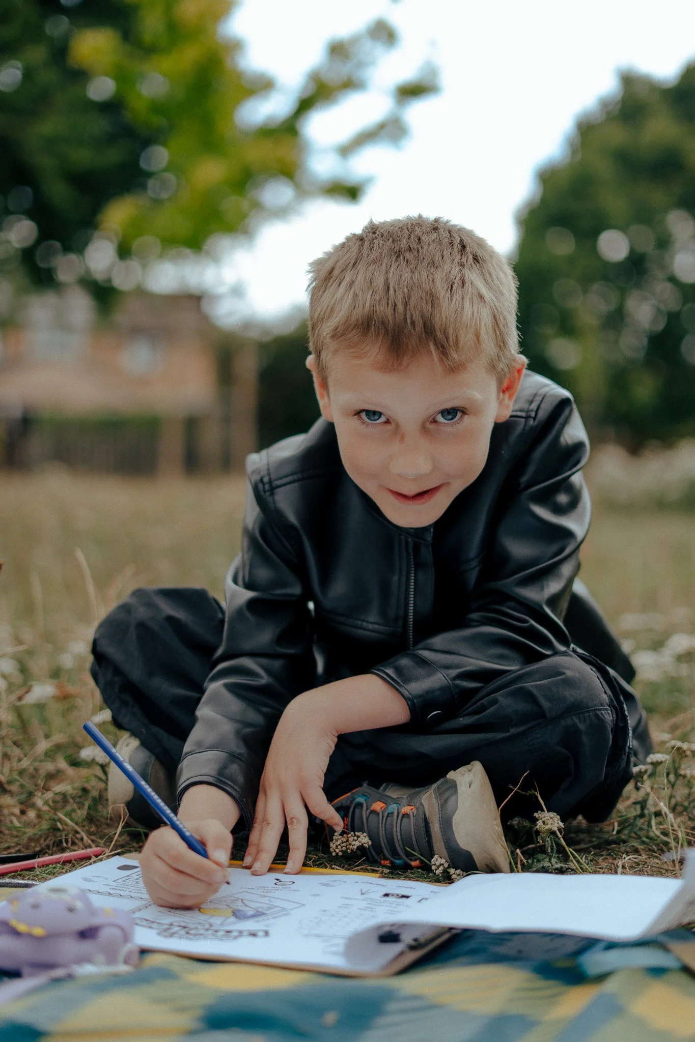 A young boy with blonde hair and blue eyes, wearing a black leather jacket and black pants, is sitting on the ground outdoors, coloring in a coloring book with a blue crayon. There are some toys and a blanket nearby, and the background has trees and a house, indicating a park or backyard setting.