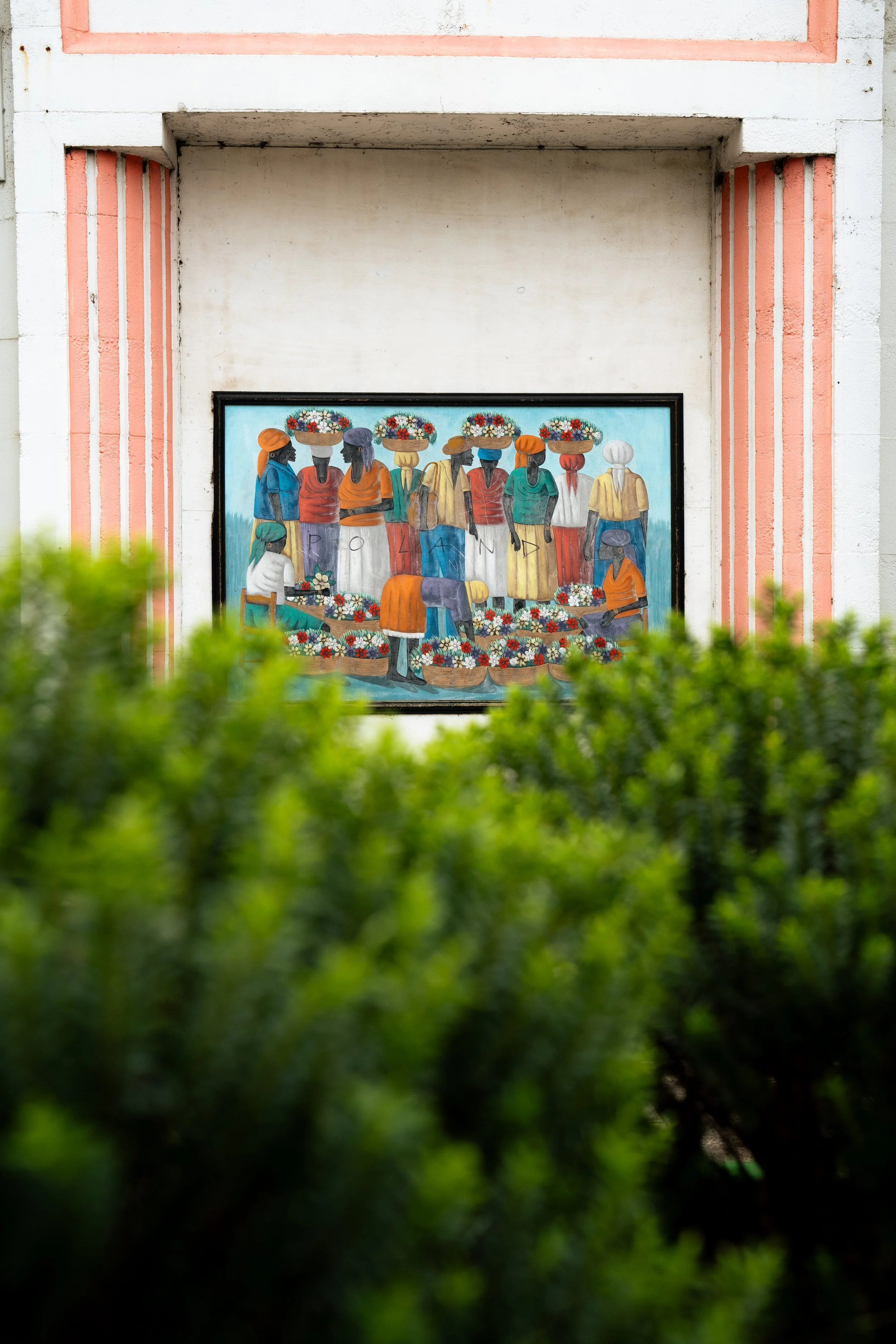 A colorful painting of people in traditional clothing engaged in a flower market, framed and mounted on a wall with pink accents, partially obscured by green shrubbery in the foreground.