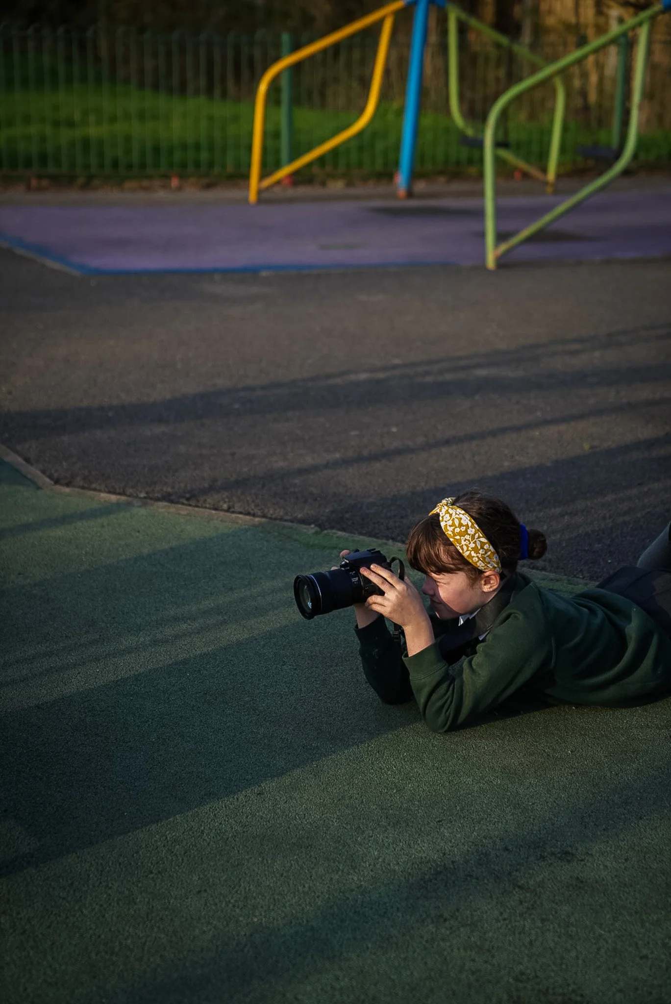 A girl lying on a green surface, with a camera, aiming to take a photo at a child's playground in the background.