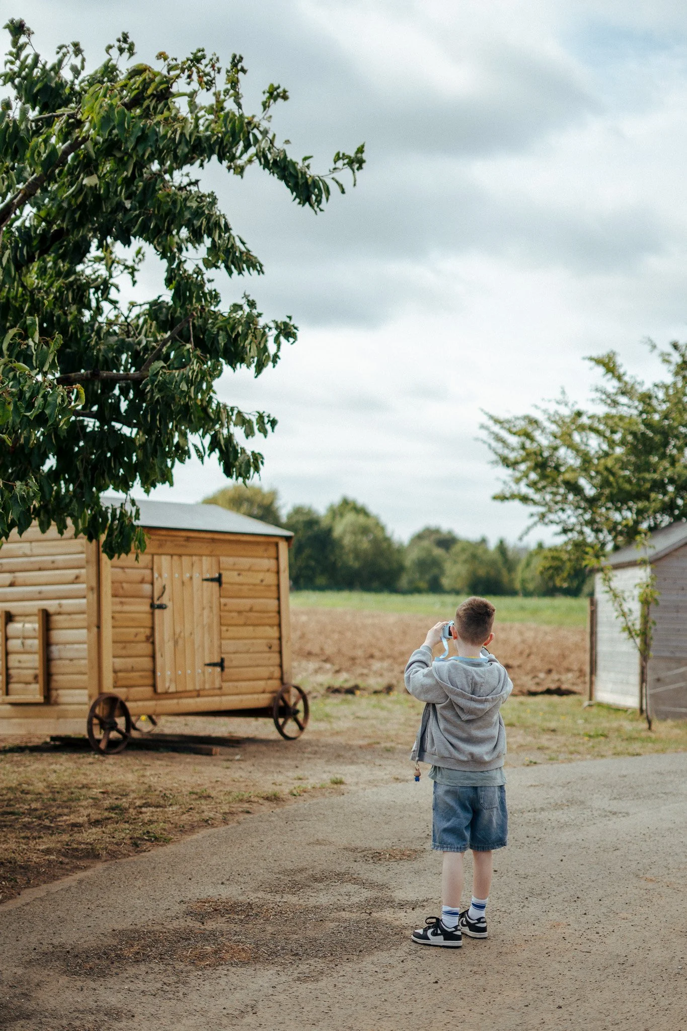 A young boy in a gray hoodie and denim shorts stands on a gravel path outdoors, taking a photo with a device, with a wooden shed on wheels and a field in the background under a cloudy sky.