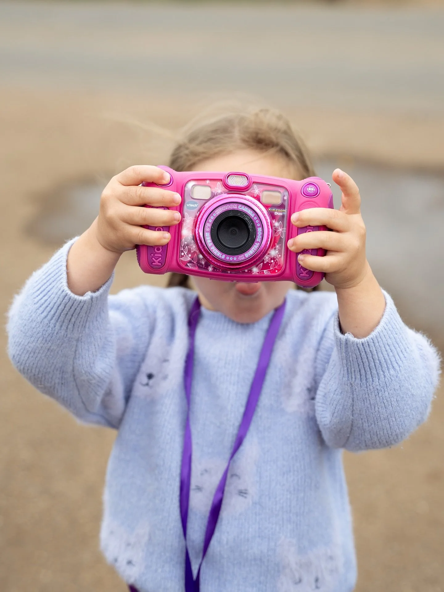 A young girl in a gray sweater holding a pink toy camera, taking a picture outdoors with a blurred background of a beach or dirt path.