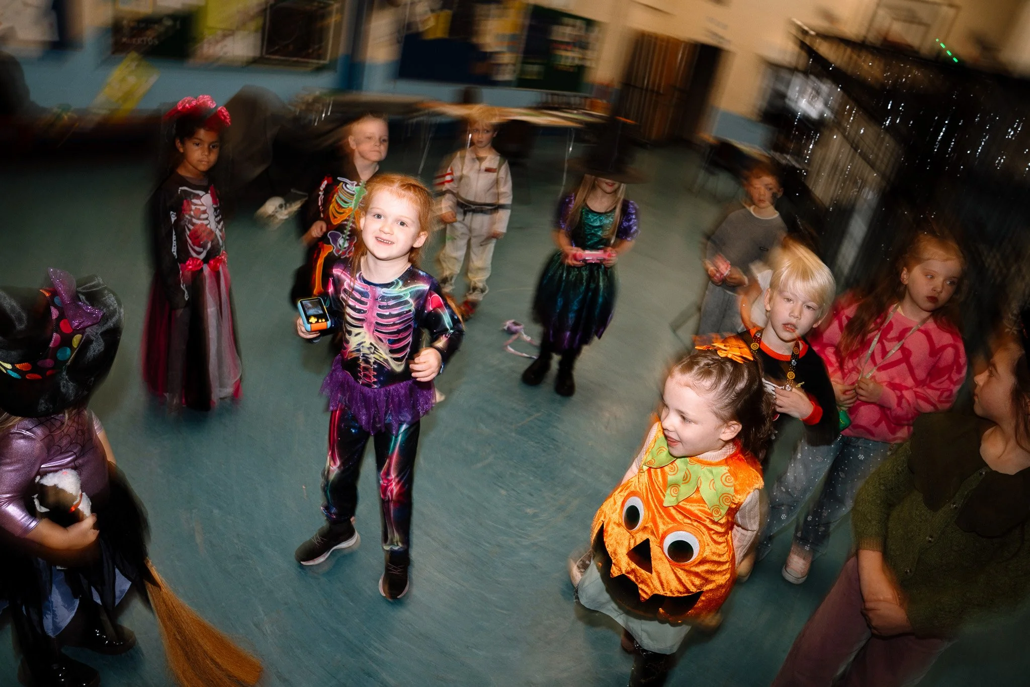 Children in Halloween costumes gathered in a room, some holding small toys or candy, with a girl dressed as a pumpkin in the foreground, looking up and smiling.