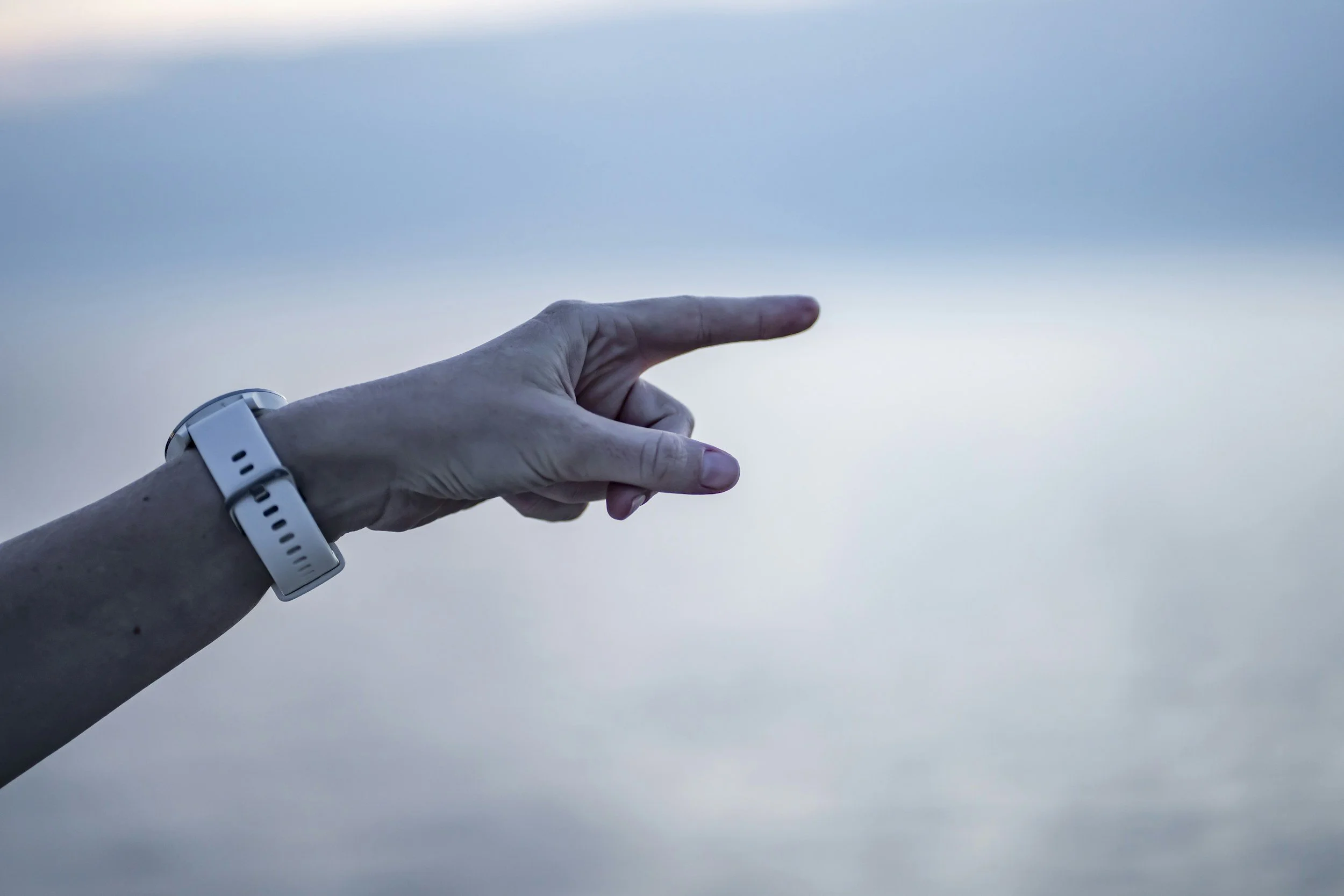 A person's hand pointing towards a light blue sky with thin clouds, wearing a watch with a white band.