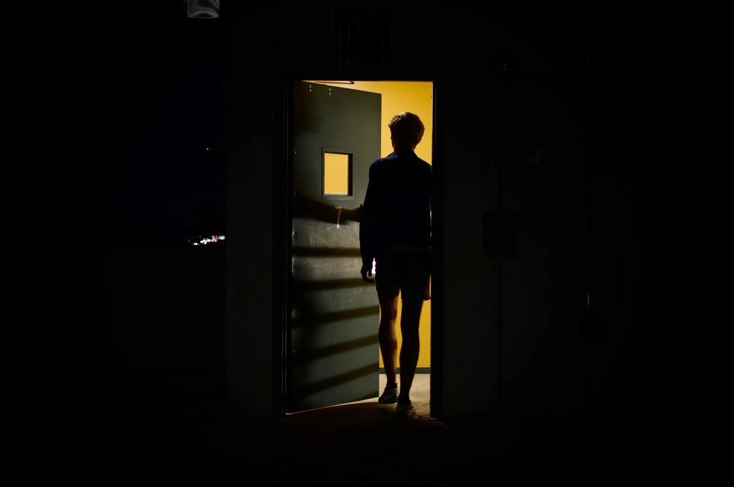 A person is walking through a doorway from darkness into a brightly lit room. The person's back is to the camera, and they are wearing shorts and sneakers.