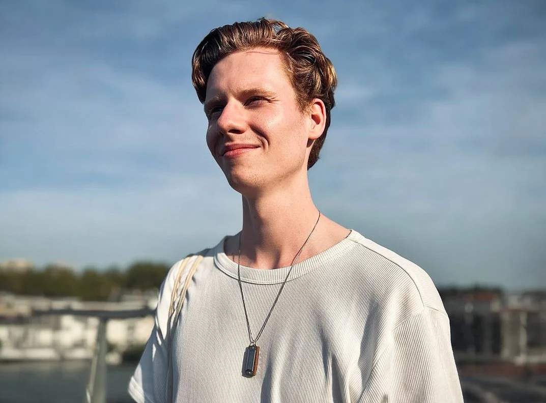 Portret van een jonge man met kort, stijlvol gekapt haar, zittend buiten onder een blauwe hemel, met een witte trui en een ketting met een hanger.