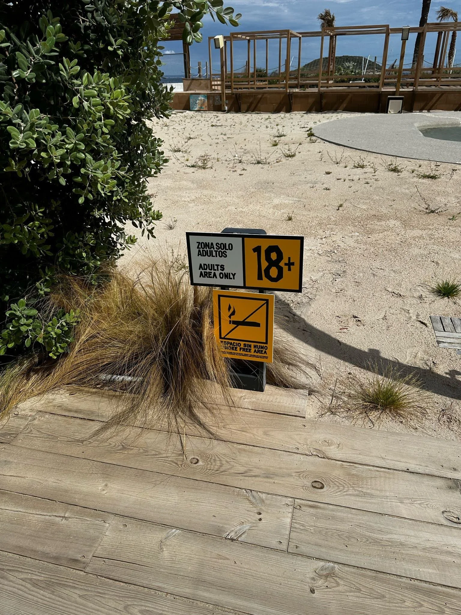 Señal que indica que solo adultos mayores de 18 años pueden acceder a la zona, en una área de playa con arena, plantas y estructura de madera.