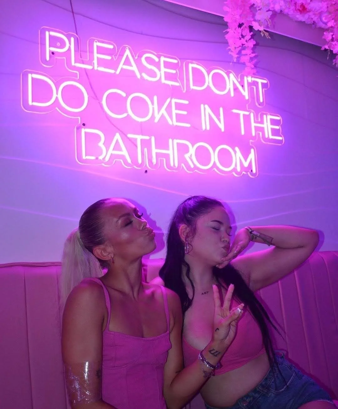 Dos mujeres sentadas en un banco en un lugar con iluminación rosada y un letrero de neón que dice 'Por favor no bebamos en el baño'.