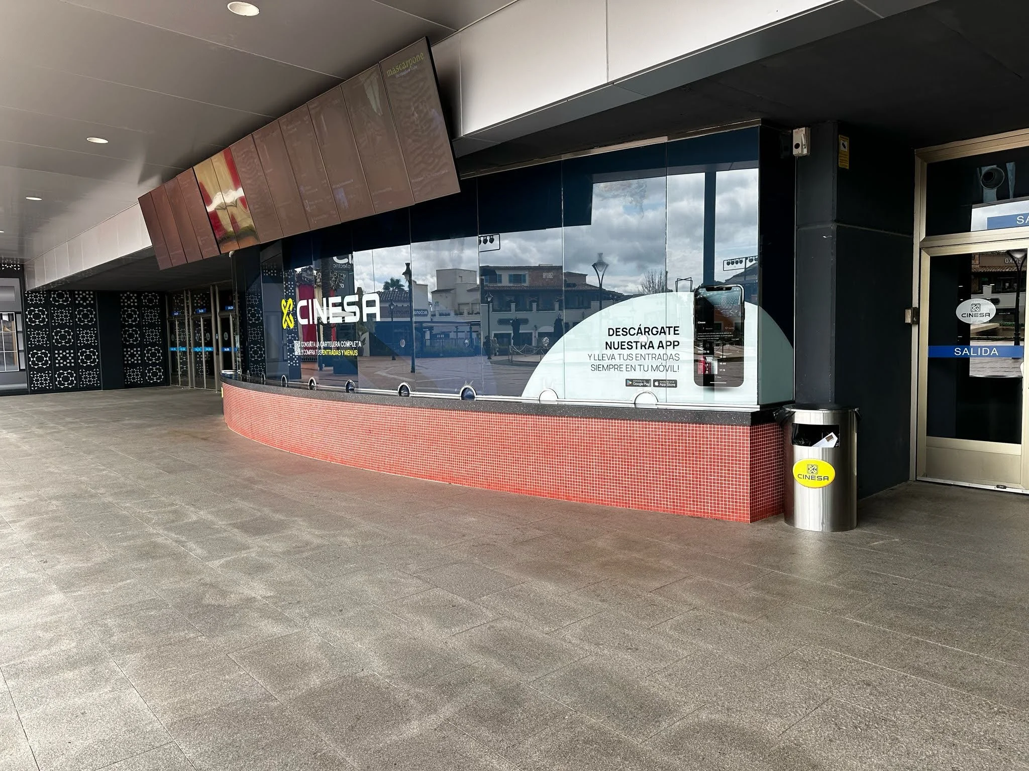 Mostrador de taquilla del cine CINESA con cartel que invita a descargar la app y llevar entradas en el móvil. Entrada y salida en la estación de cine con piso de baldosas grises y ventanas que reflejan el cielo y edificios.