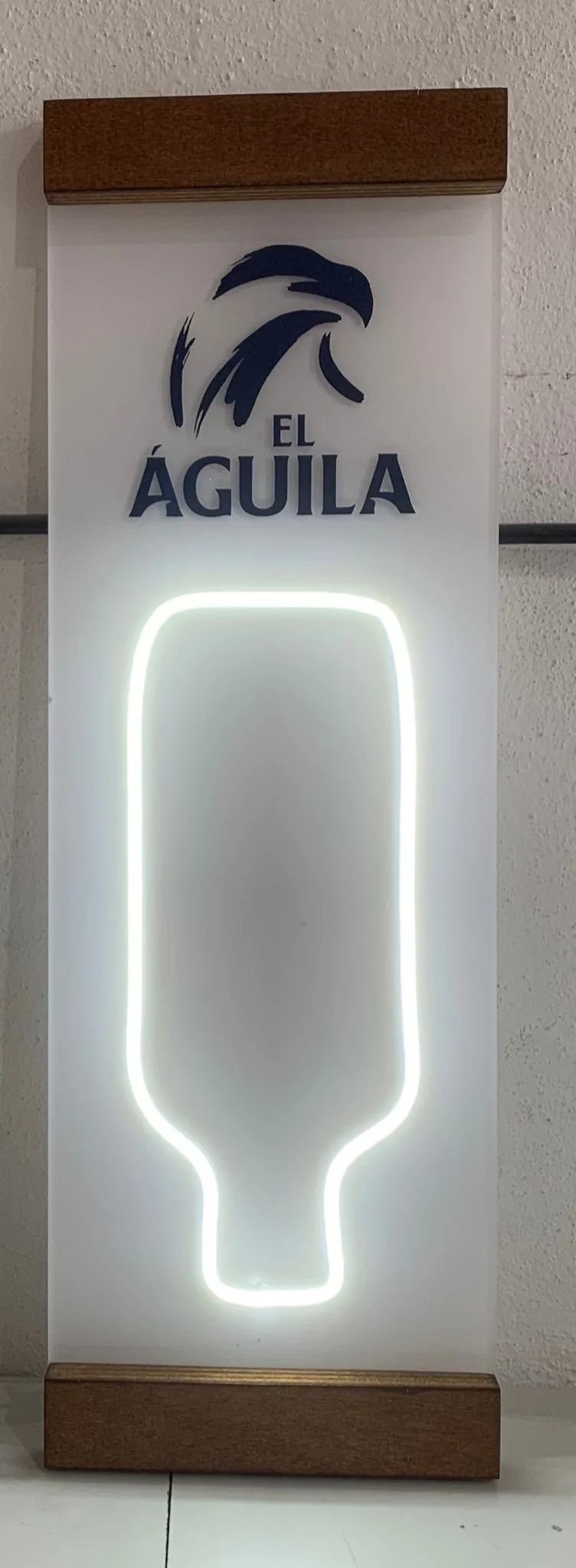 Cartel con un logotipo de un águila y las palabras "El Águila" en color azul, con un diseño de símbolo de copa iluminado en línea de neón blanca debajo.