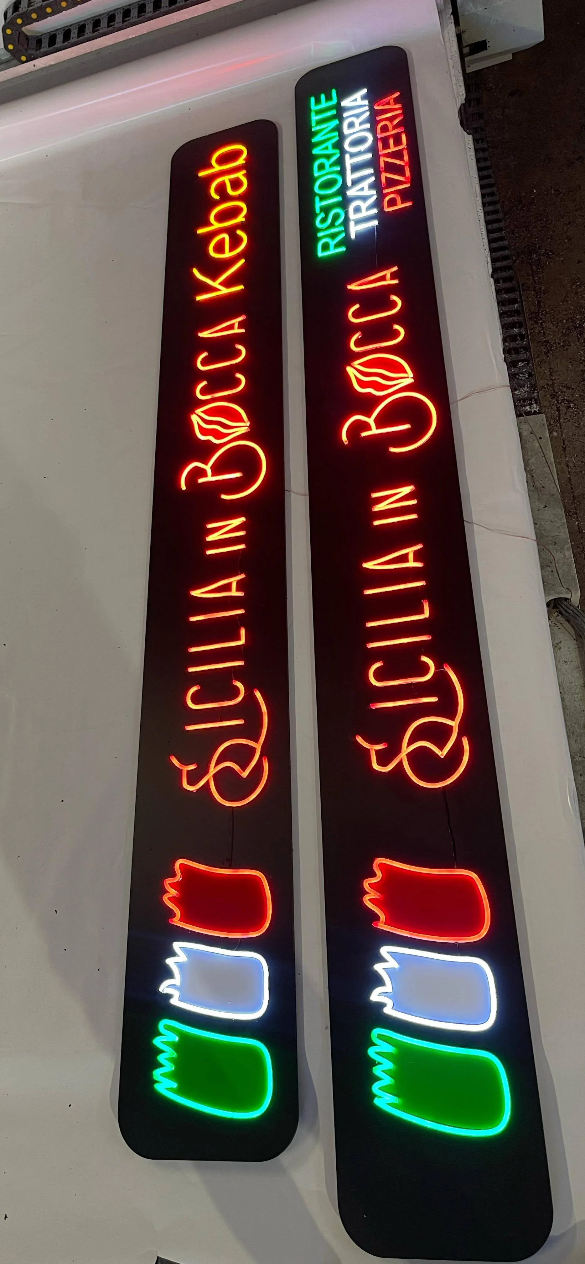 Carteles de neón en español que anuncian una cafetería, un restaurante trattoria y una pizzería con colores rojo, verde, blanco y azul, y símbolos de pizza y una mano haciendo gesto de paz.