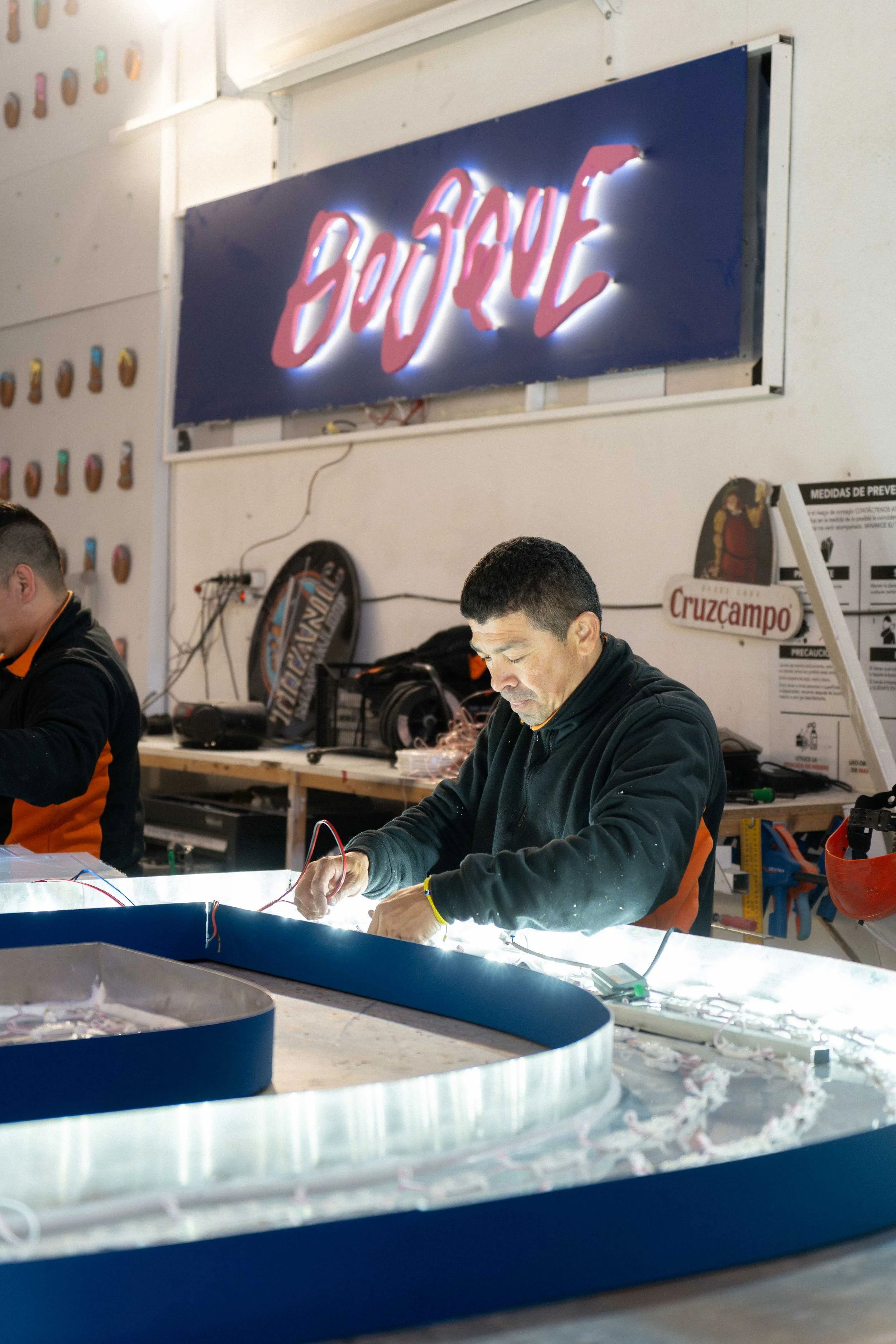 Hombre trabajando en una línea de producción con luces LED y un letrero de neón que dice 'Bourré' en el fondo.