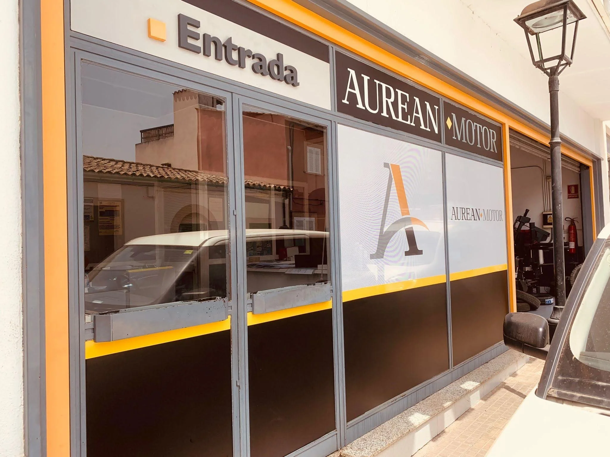Fachada de un taller mecánico llamado Aurean Motor, con un cartel grande, ventanas de vidrio y una lámpara en la esquina. Hay un coche y un camión viejos en el exterior.