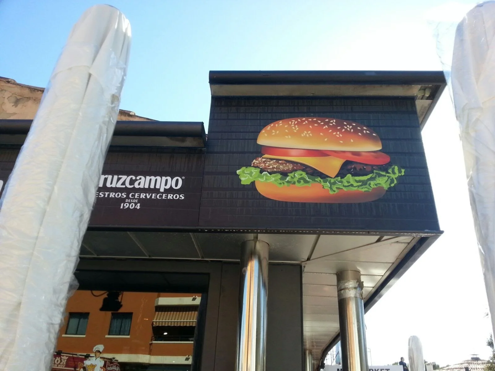 Cartel con imagen de una hamburguesa con queso, lechuga, tomate, cebolla y pan con semillas.