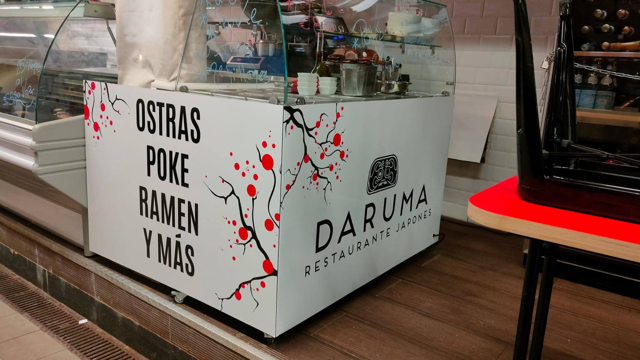 Mostrador de restaurante japonés con cartel que dice 'Ostras, Poke, Ramen y Más', decorado con ramas y puntos rojos, y un refrigerador de helados a la izquierda y una parrilla eléctrica a la derecha.