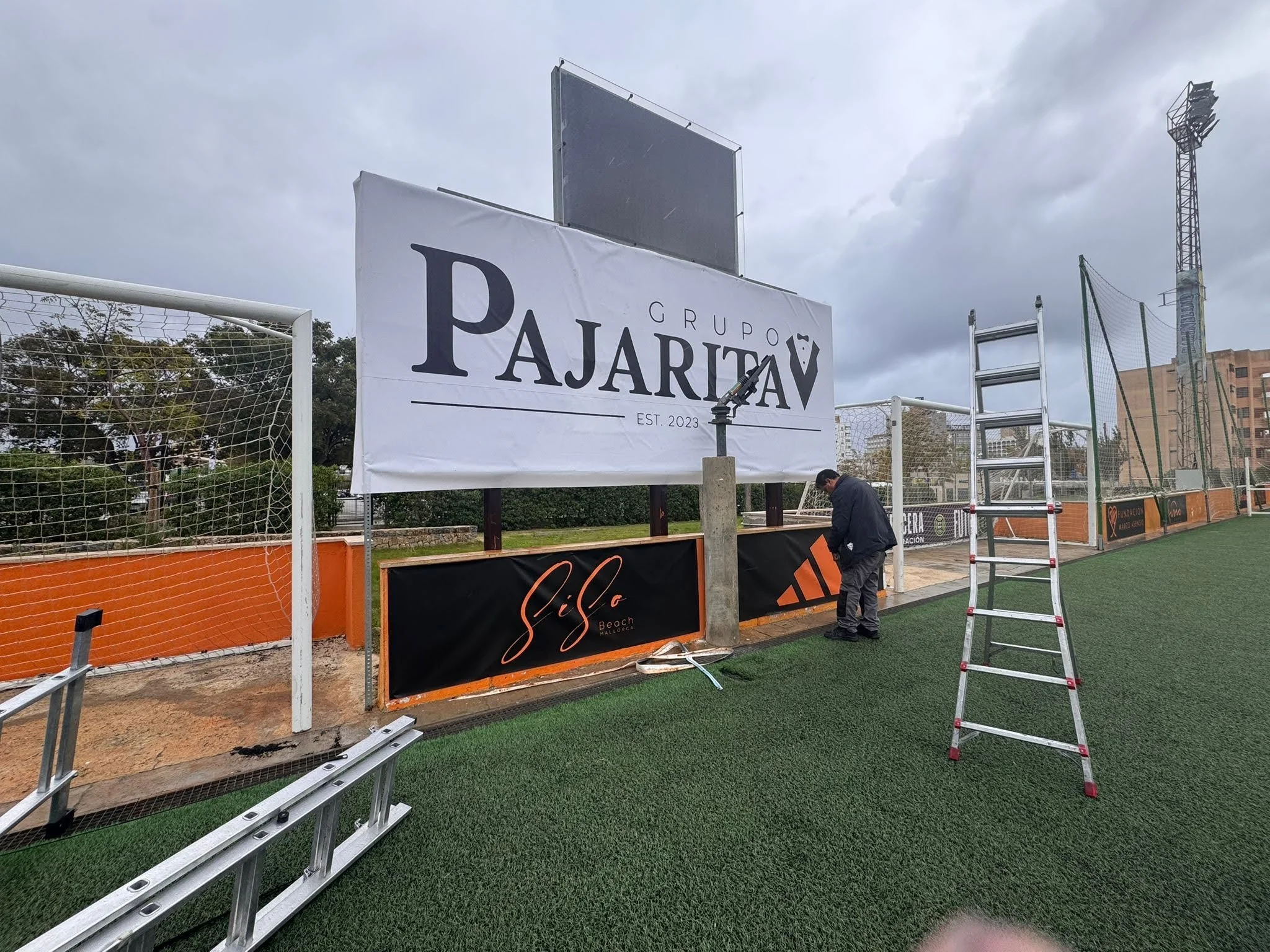 Campo de fútbol en construcción con vallas y anuncios publicitarios, y una persona trabajando en los detalles.