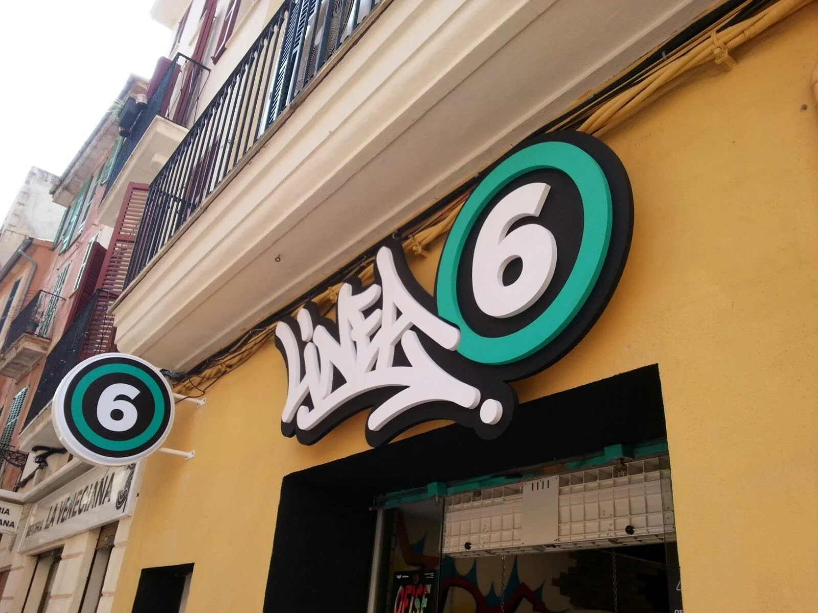 Cartel con número 6 en la fachada de un establecimiento, decorado con letra estilo graffiti, en un edificio de plusieurs pisos con balcones y ventanas con barandales de metal.