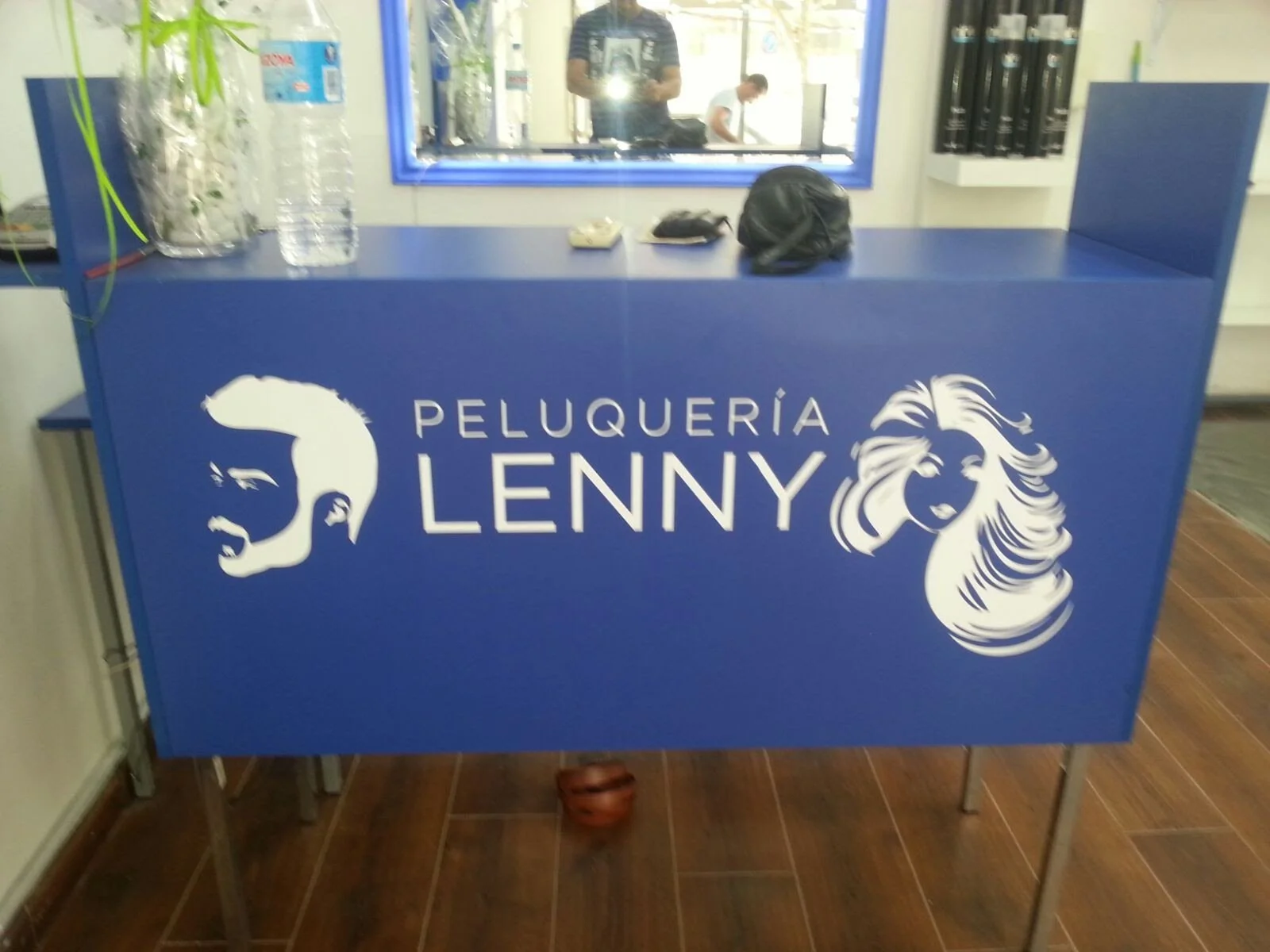 Mostrador de peluquería azul con el logotipo y nombre Peluquería Lenny, con ilustraciones de un hombre y una mujer en blanco, en el fondo un espejo y algunos objetos.