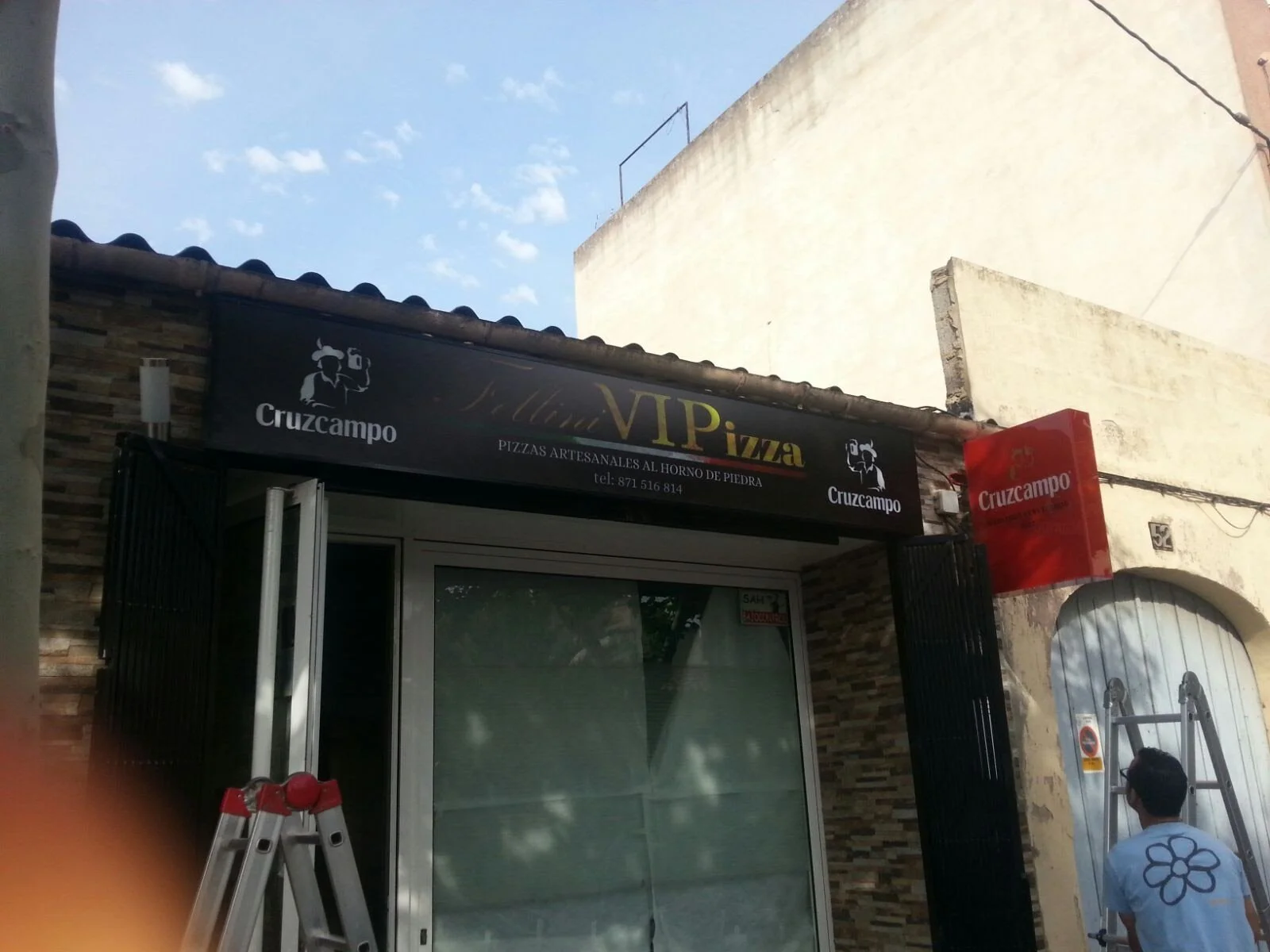 Cartel de una pizzería llamada 'VIP Pizza' que ofrece pizzas artesanales al horno de piedra. Hay una persona trabajando en la puerta, un andamio y un letrero rojo con la misma marca 'Cruzcampo'.