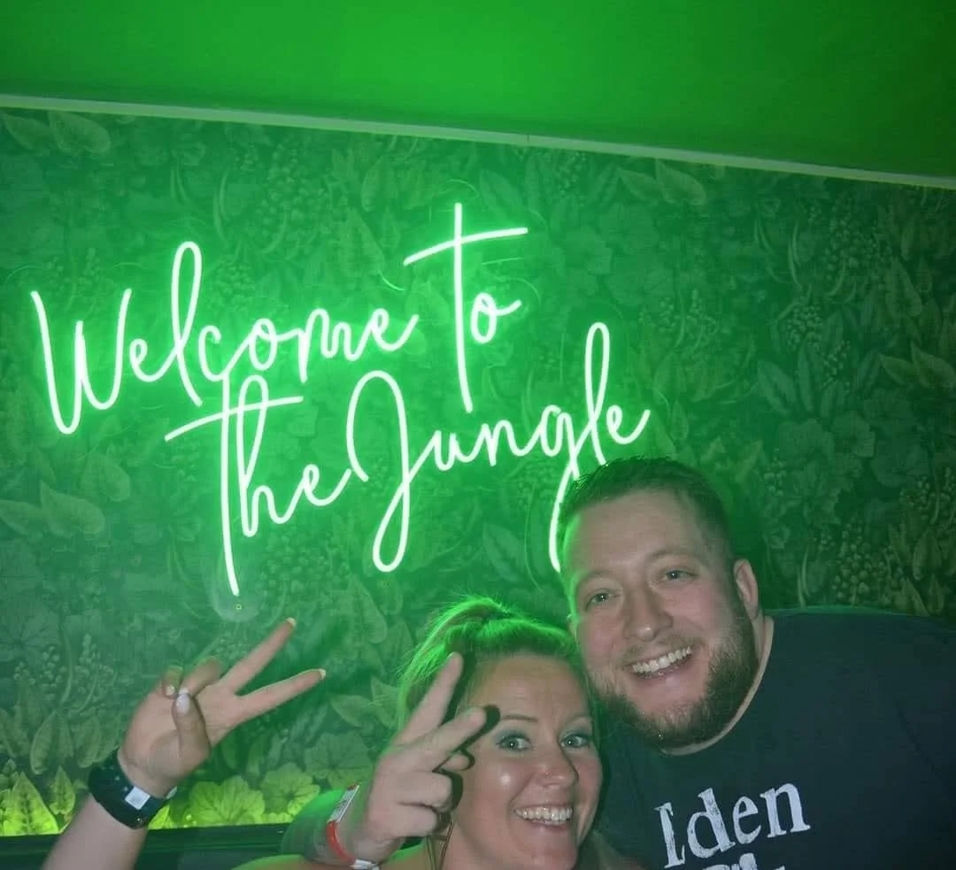Una mujer y un hombre sonriendo en un lugar con decoración de jungla y un cartel de neón que dice "Welcome to the Jungle" en letras verdes, haciendo señal de paz con las manos.