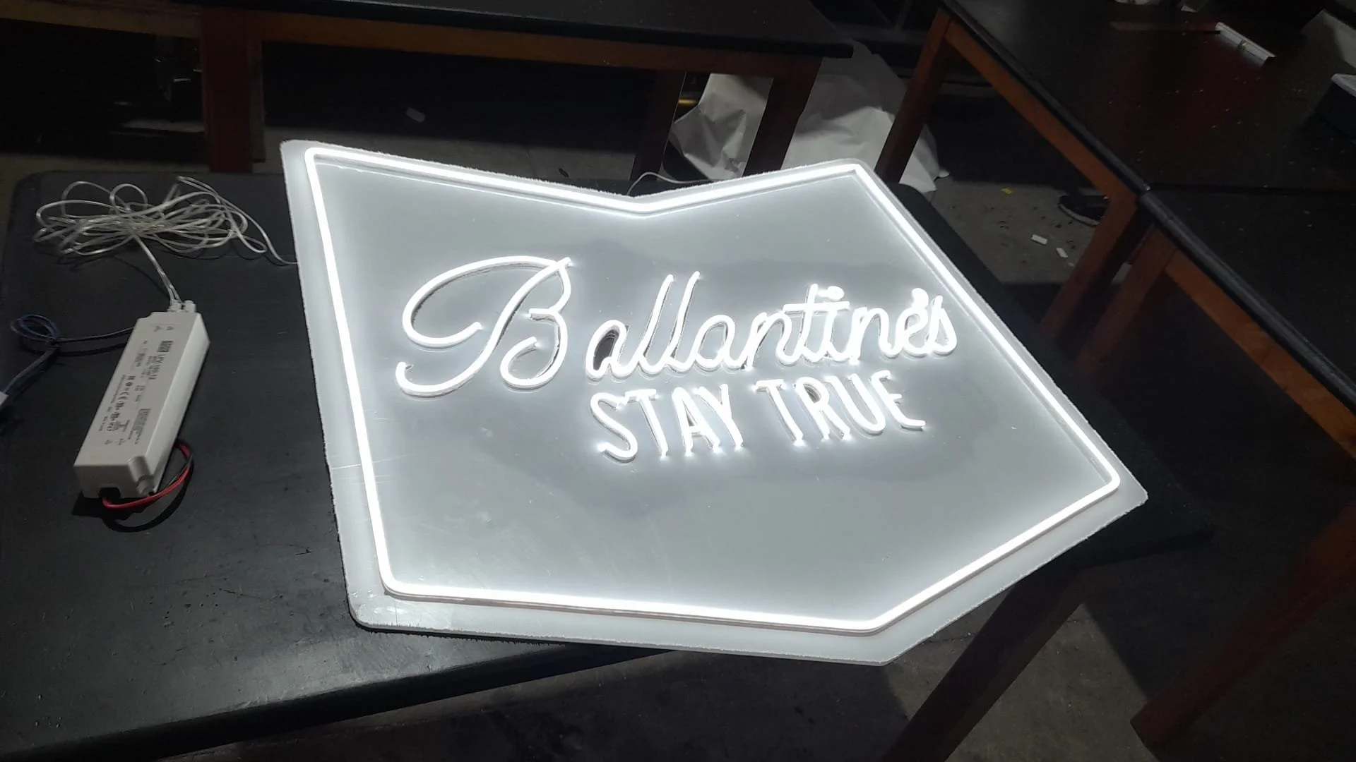 Letrero de neón que dice 'Ballantine's STAY TRUE' en letra cursiva y mayúscula sobre un fondo transparente, conectado a un transformador eléctrico, en un entorno de taller o taller artesanal.