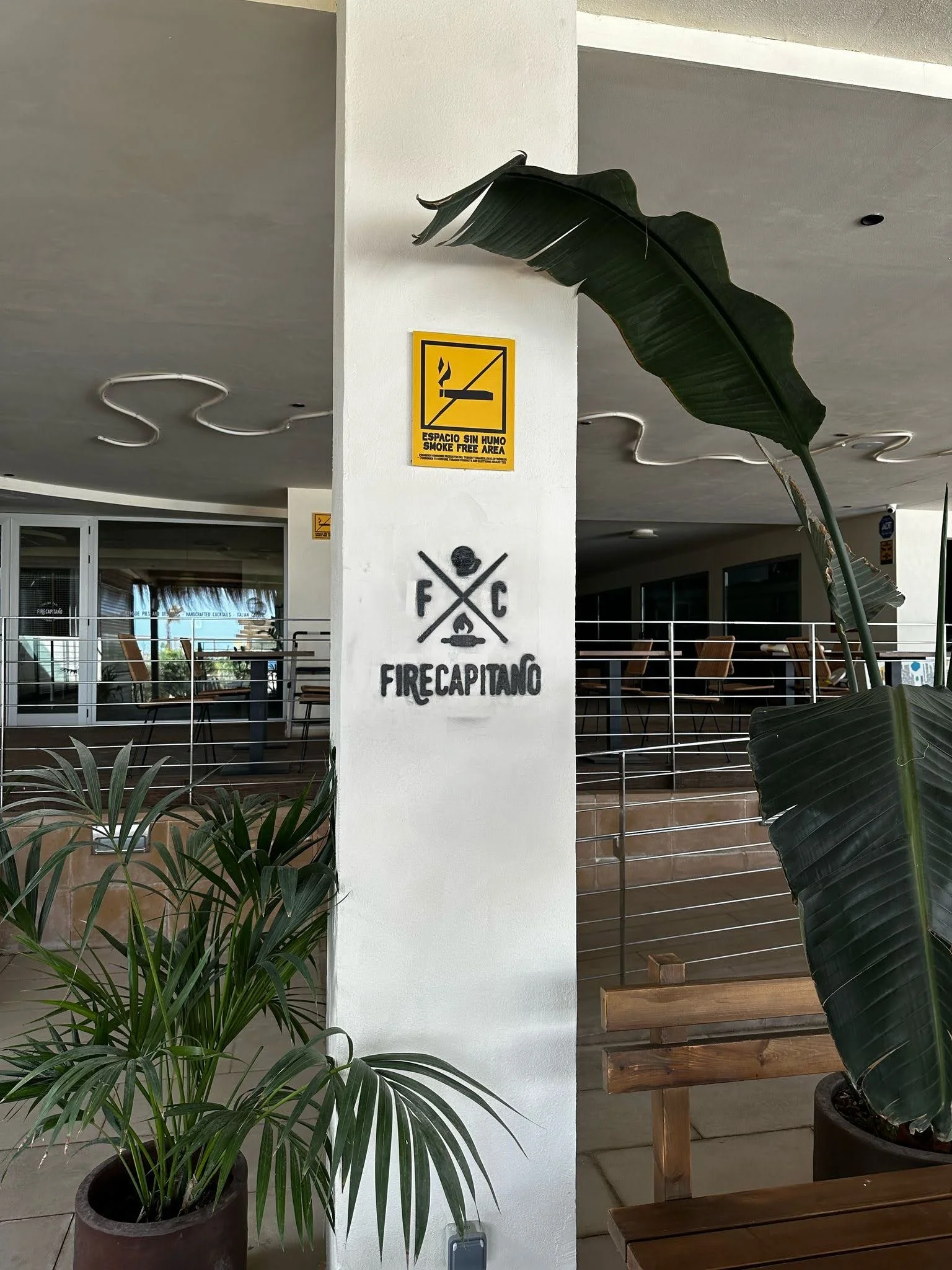 Área de descanso en un espacio interior con plantas en macetas y señal de advertencia de área sin humo en la pared, con mesas y sillas de madera.
