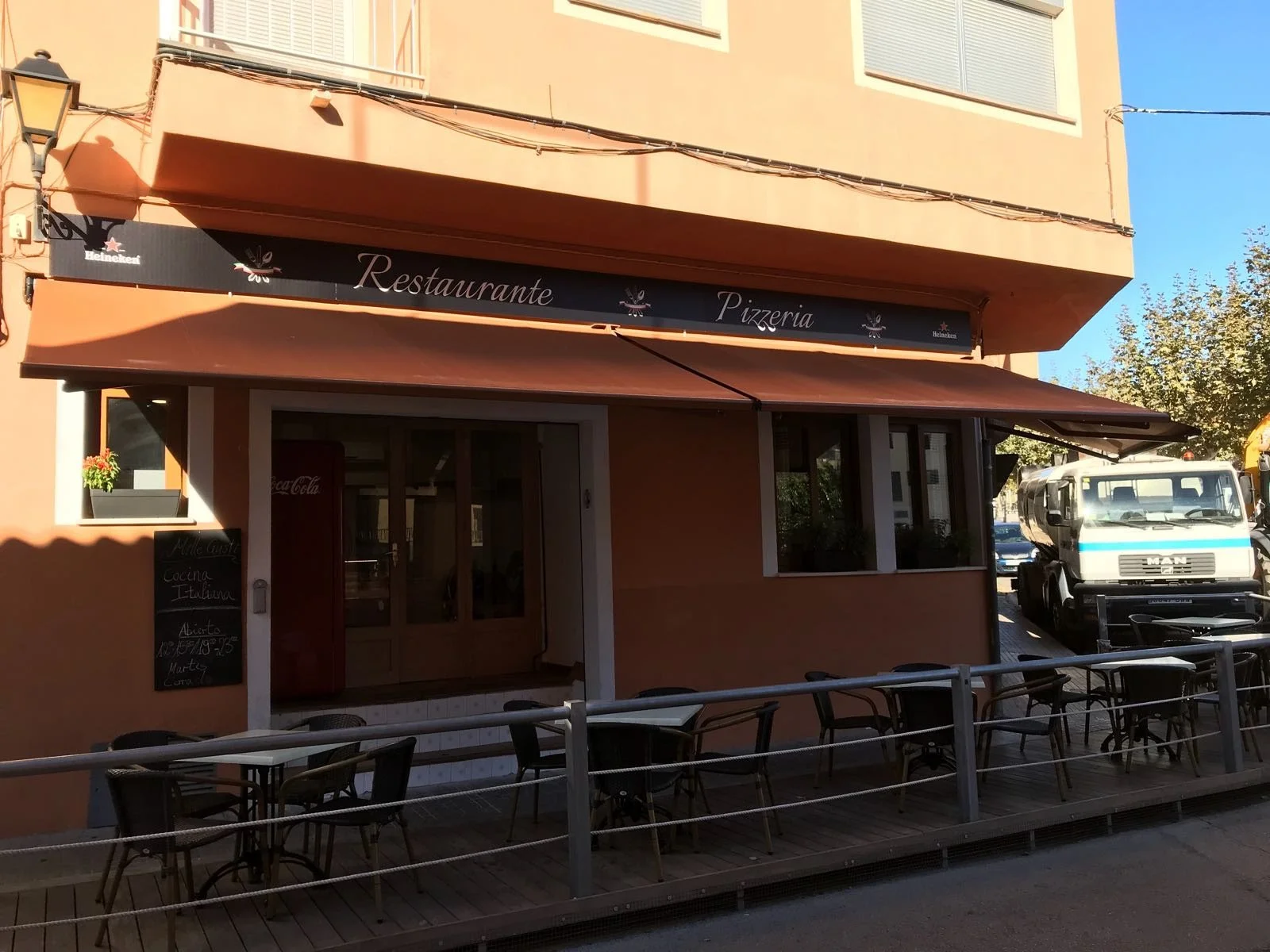 Fachada de un restaurante y pizzería con terraza abierta, toldos de color marrón, y un cartel con horarios, en una calle con árboles y un camión blanco y azul estacionado.