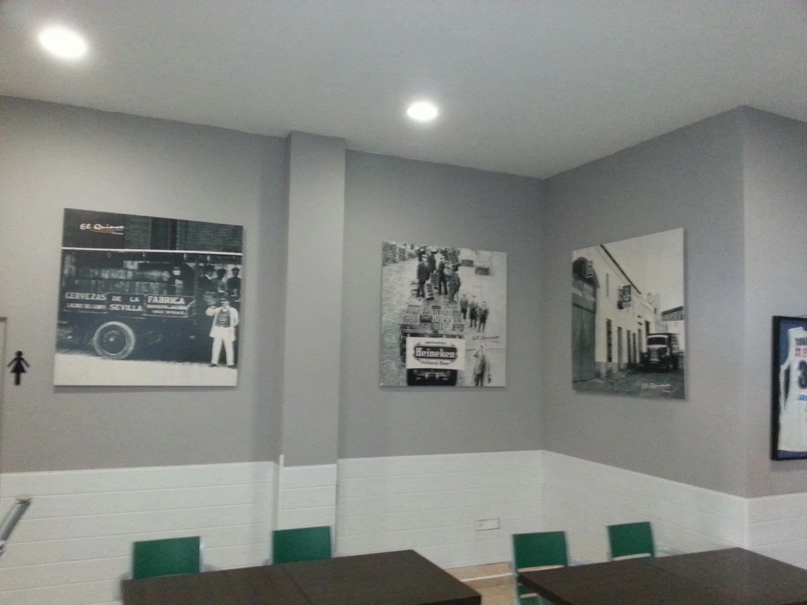 Cuatro cuadros en una pared gris con sillas verdes y mesas de madera en primer plano, en ambiente de cafetería o restaurante.