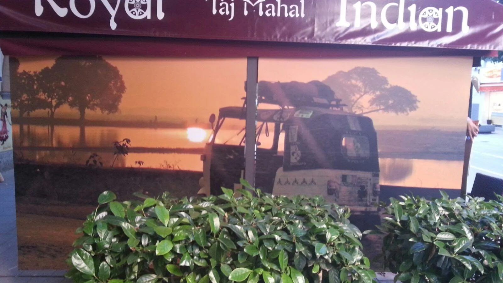 Cartel publicitario con imagen de un paisaje al amanecer, con un río y árboles, y un vehículo tipo tuk-tuk en primer plano.