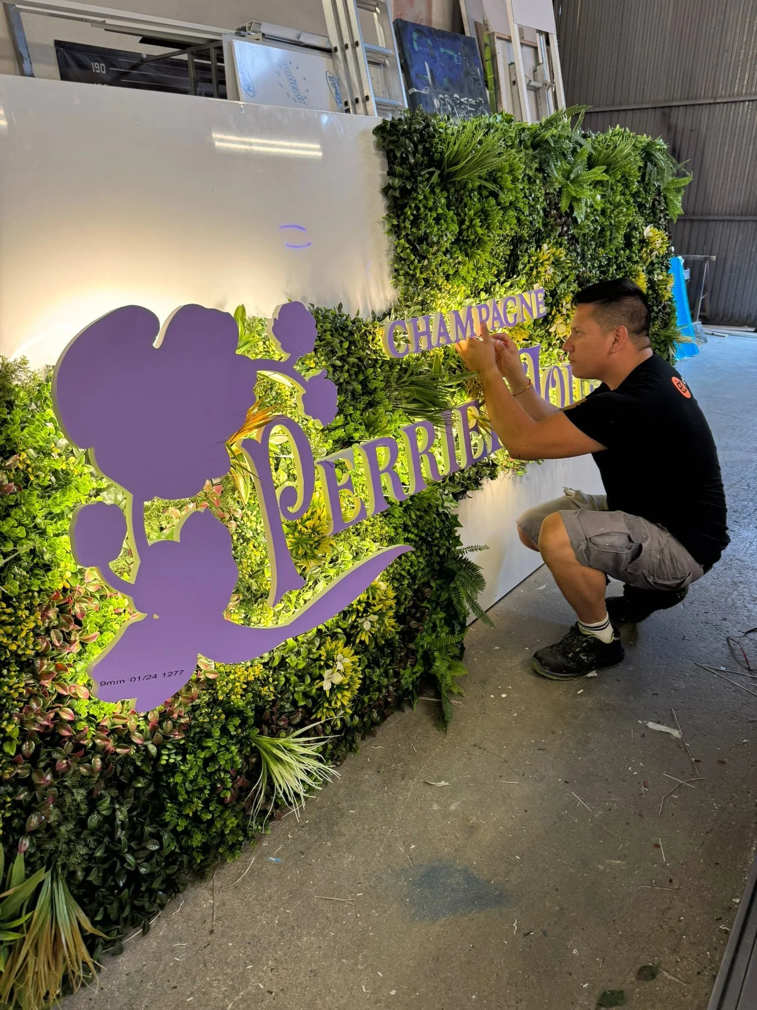 Joven decorando un arreglo de flores con letras moradas que dicen "Champagne" y "Derby" en un espacio industrial con paredes de metal y pisos de concreto.
