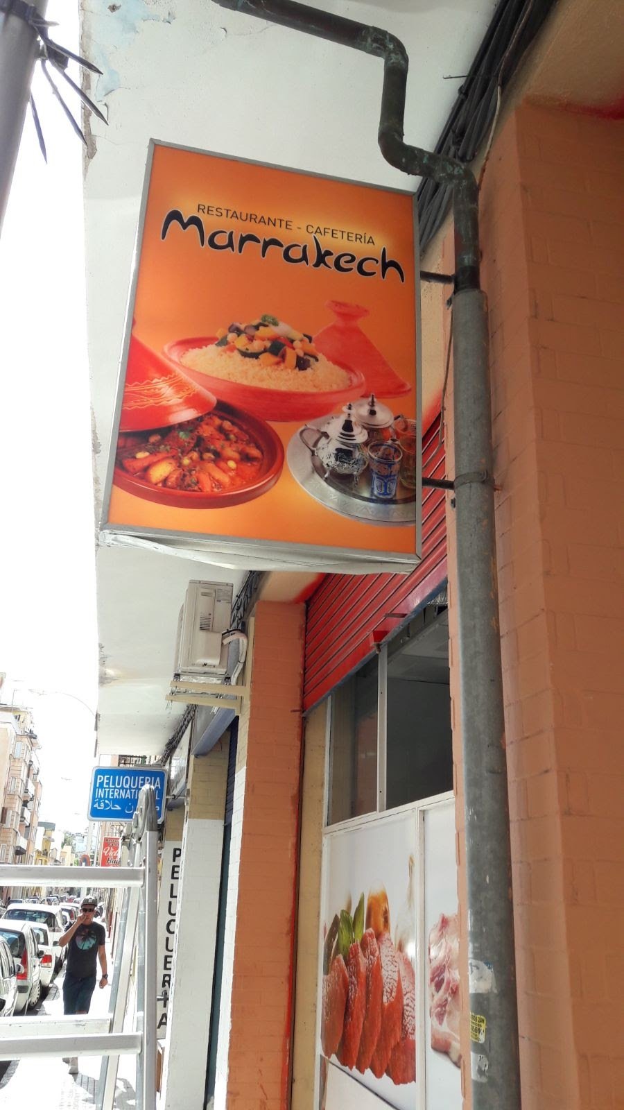 Cartel de restaurante y cafeteria 'Marrakech' con imagen de comida marroquí, incluyendo un plato de cuscús y tajín en platos de barro, en un entorno urbano.