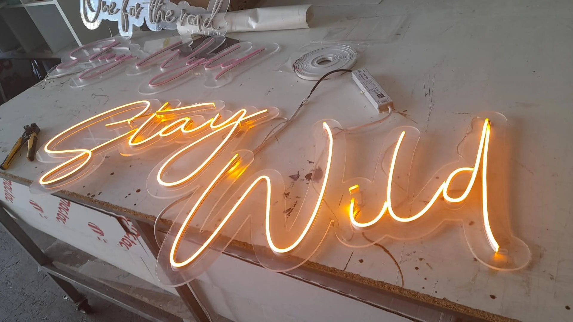 Letras de neón que dicen 'Say Wild' colocadas sobre una mesa, con herramientas y cables visibles alrededor, en un entorno de trabajo para fabricación de letreros luminosos.