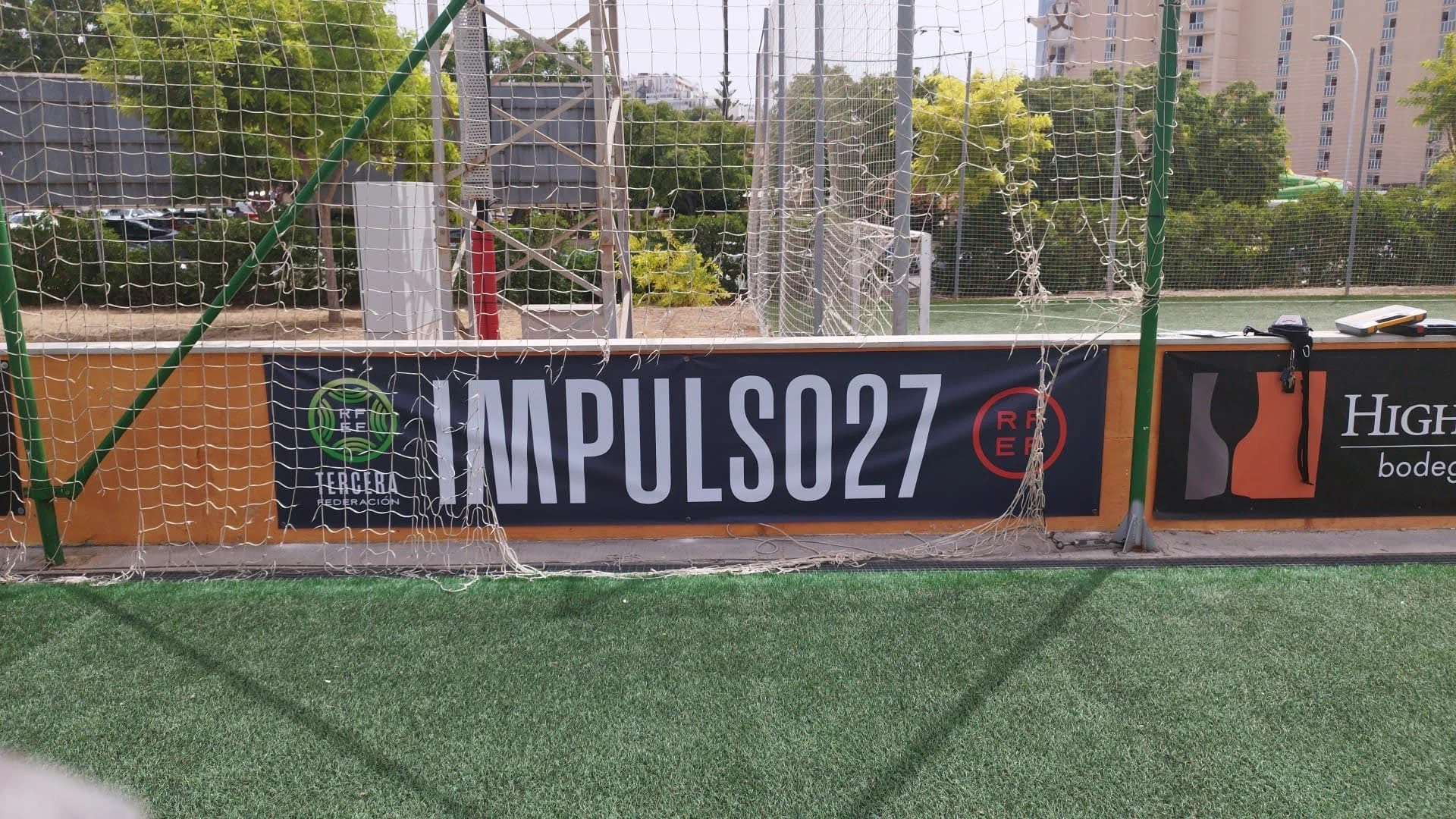 Valla de alambre en una cancha de fútbol con anuncios publicitarios, algunos árboles y edificios en el fondo.