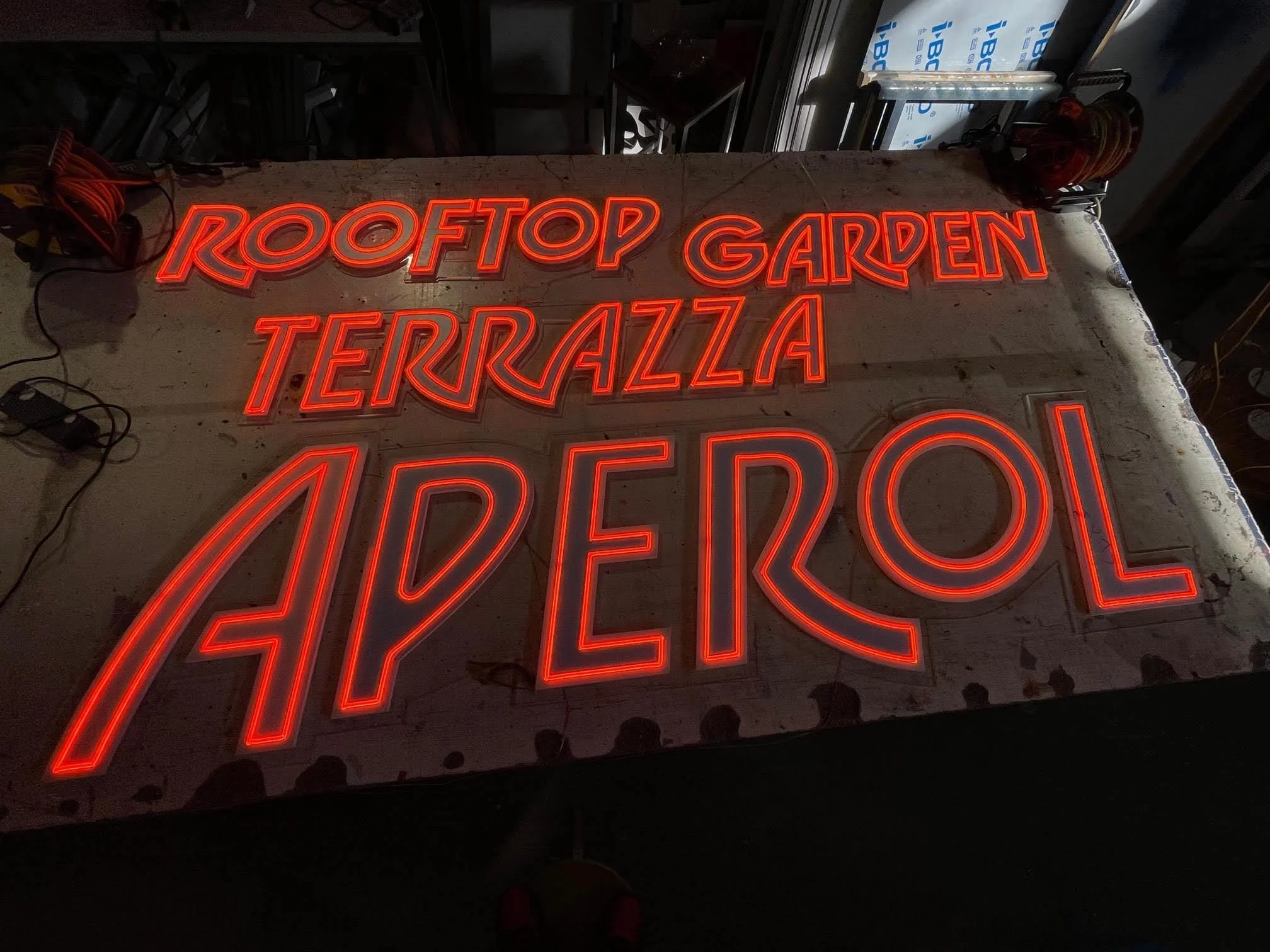 Una cartel luminoso de neón en color rojo que dice 'ROOFTOP GARDEN TERRAZZA' en un taller o almacén, rodeado de cables y herramientas.